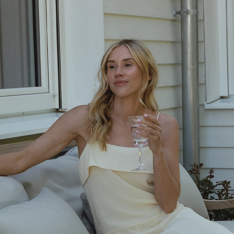 Vrouw in crèmekleurige jurk ontspant buiten met glas in hand op tuinbank