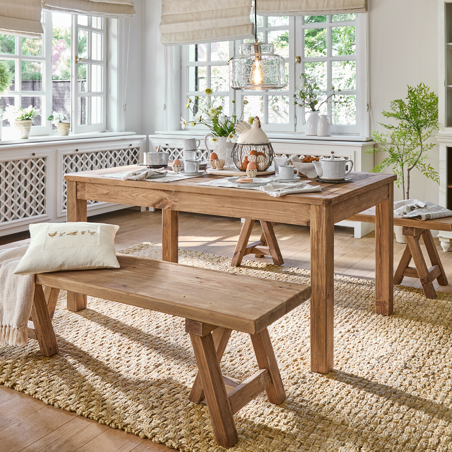 Eetkamer met houten tafel en banken op natuurlijke vloerkleed, lichte inrichting met decoraties en licht