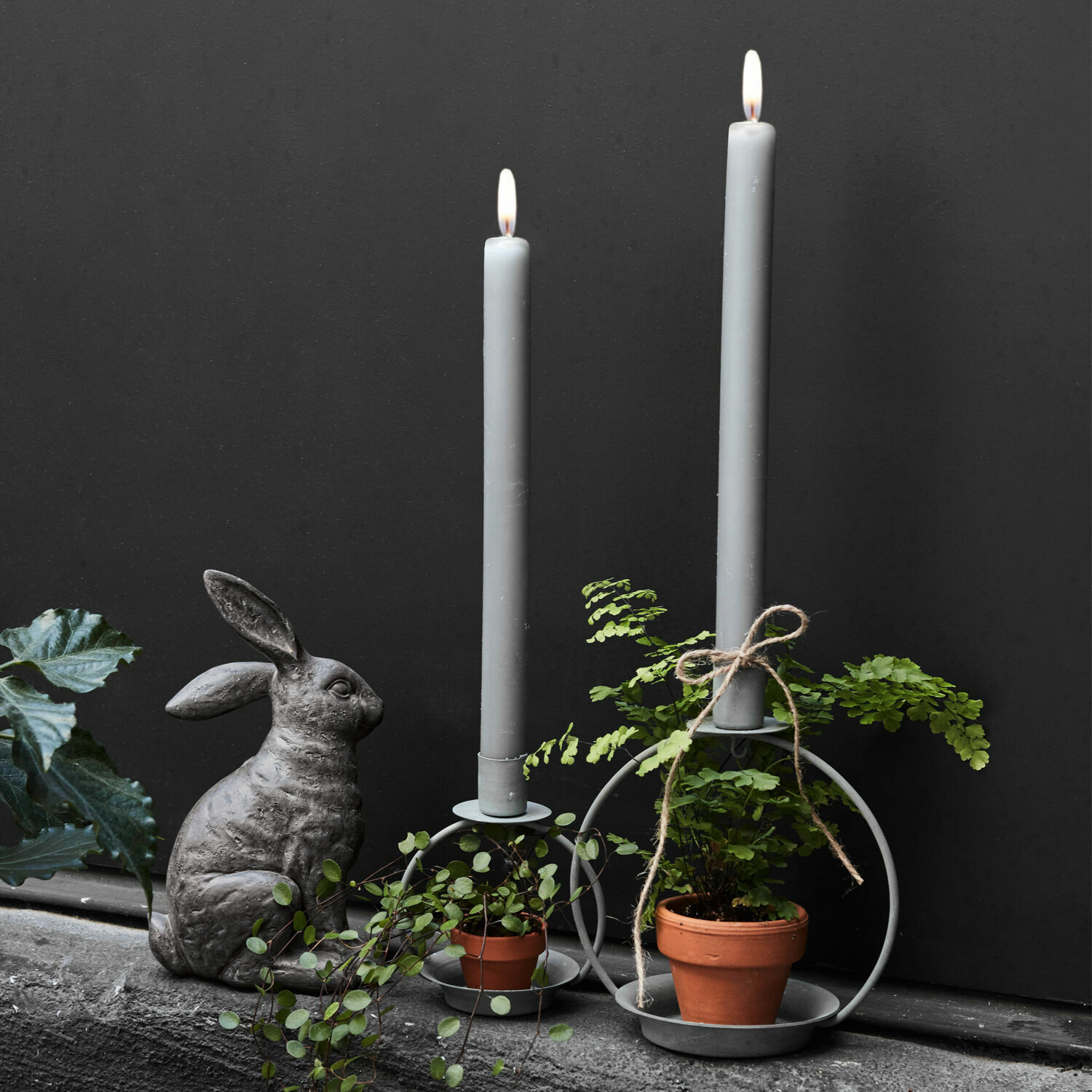 Grijze metalen kandelaren met grijze kaarsen, groene planten in potten en stenen konijn decoratie