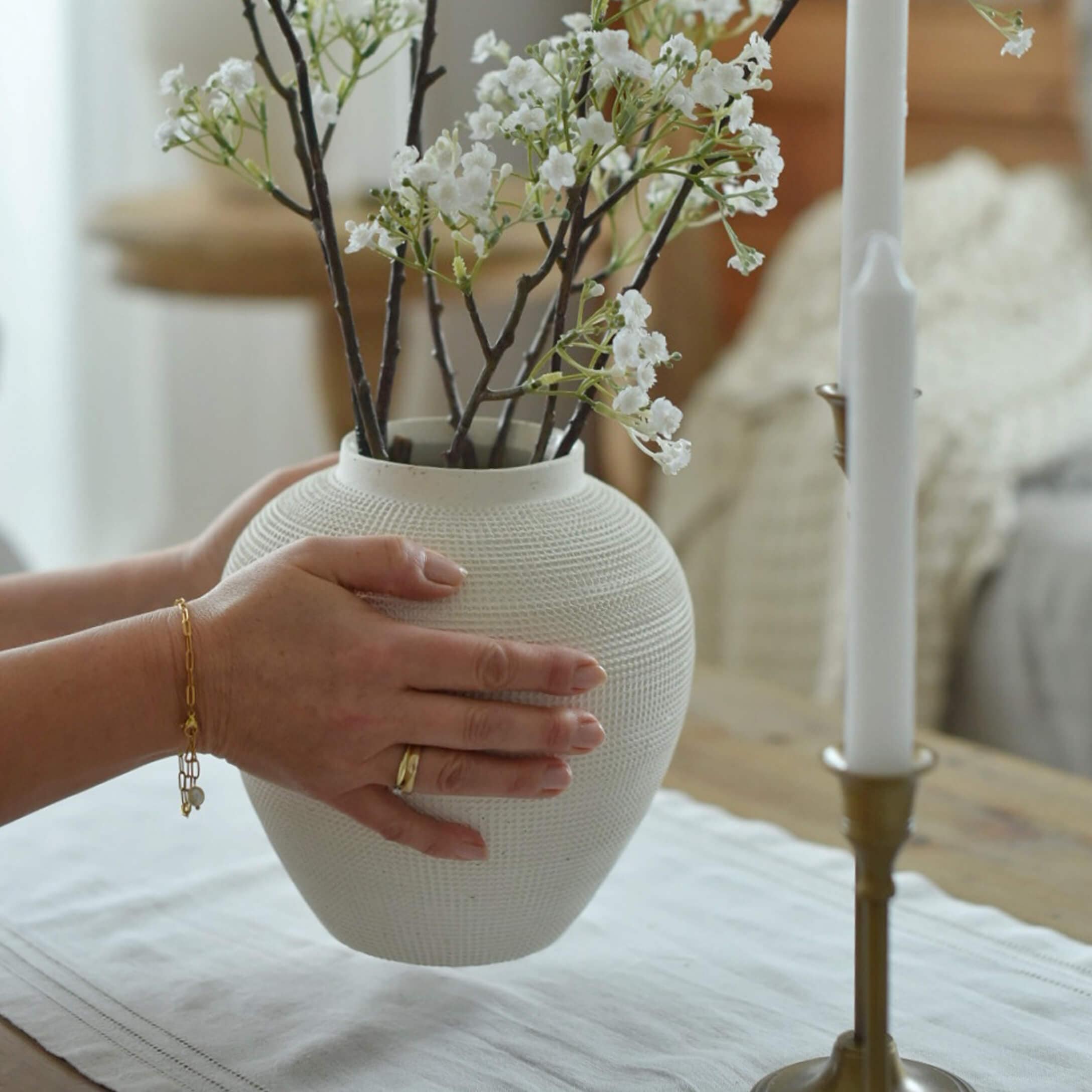 Witte keramische vaas met textuur vastgehouden door handen met takken witte bloemen op tafel