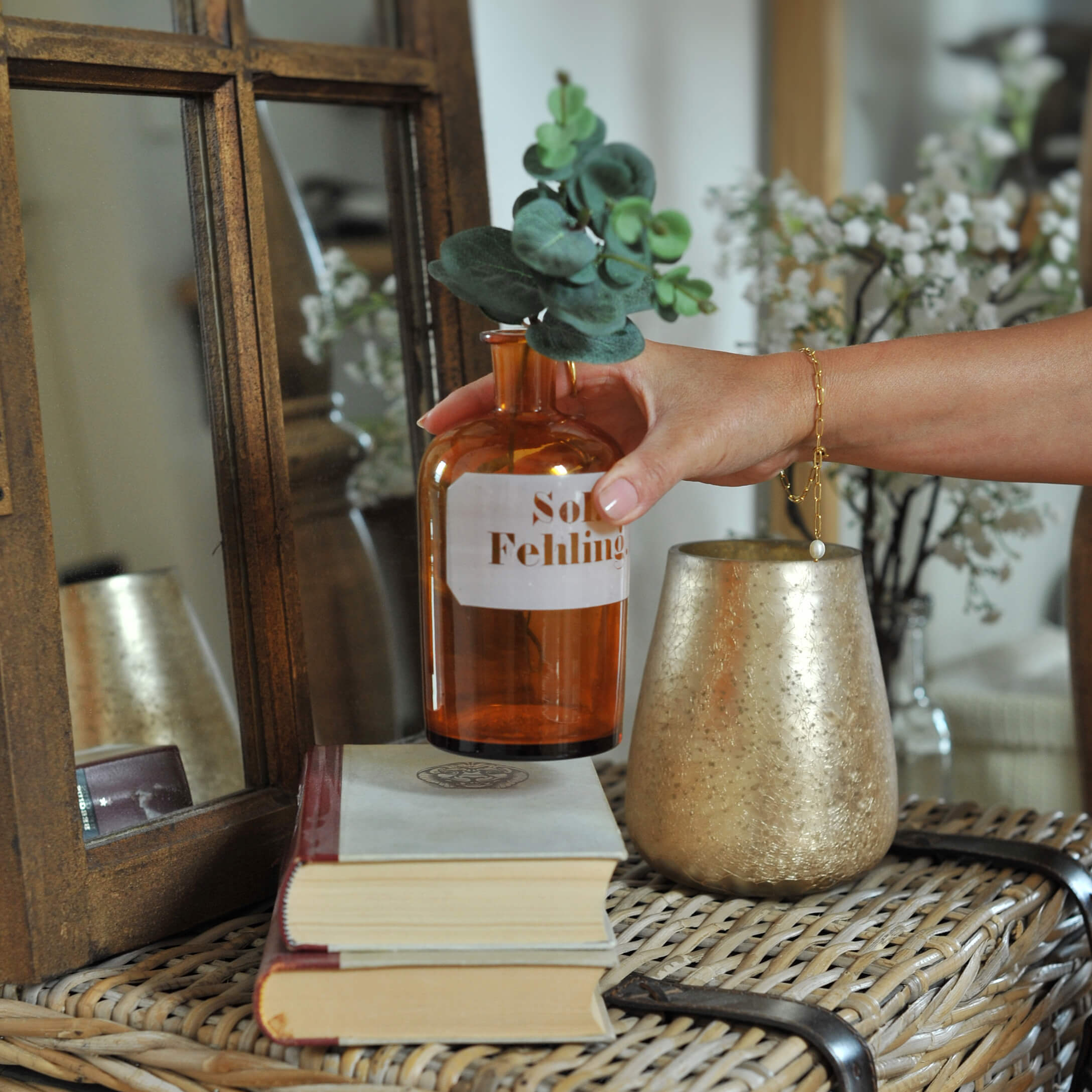 Hand houdt amberkleurige glazen fles met groene bladeren op rotan tafel met boeken en gouden kandelaar