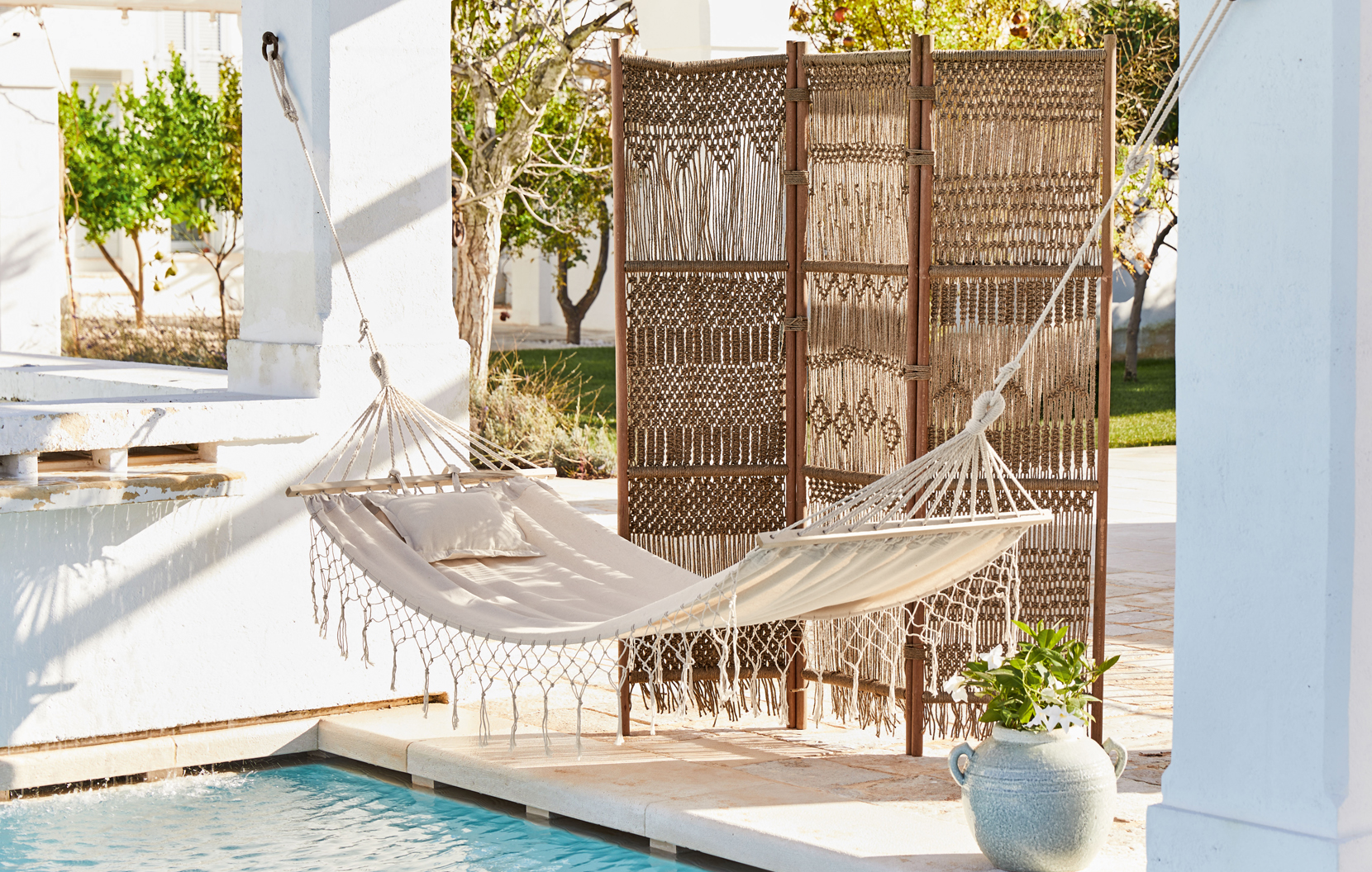 Beige stoffen hangmat met kussen en gevlochten kamerscherm op terras bij zwembad in boho stijl