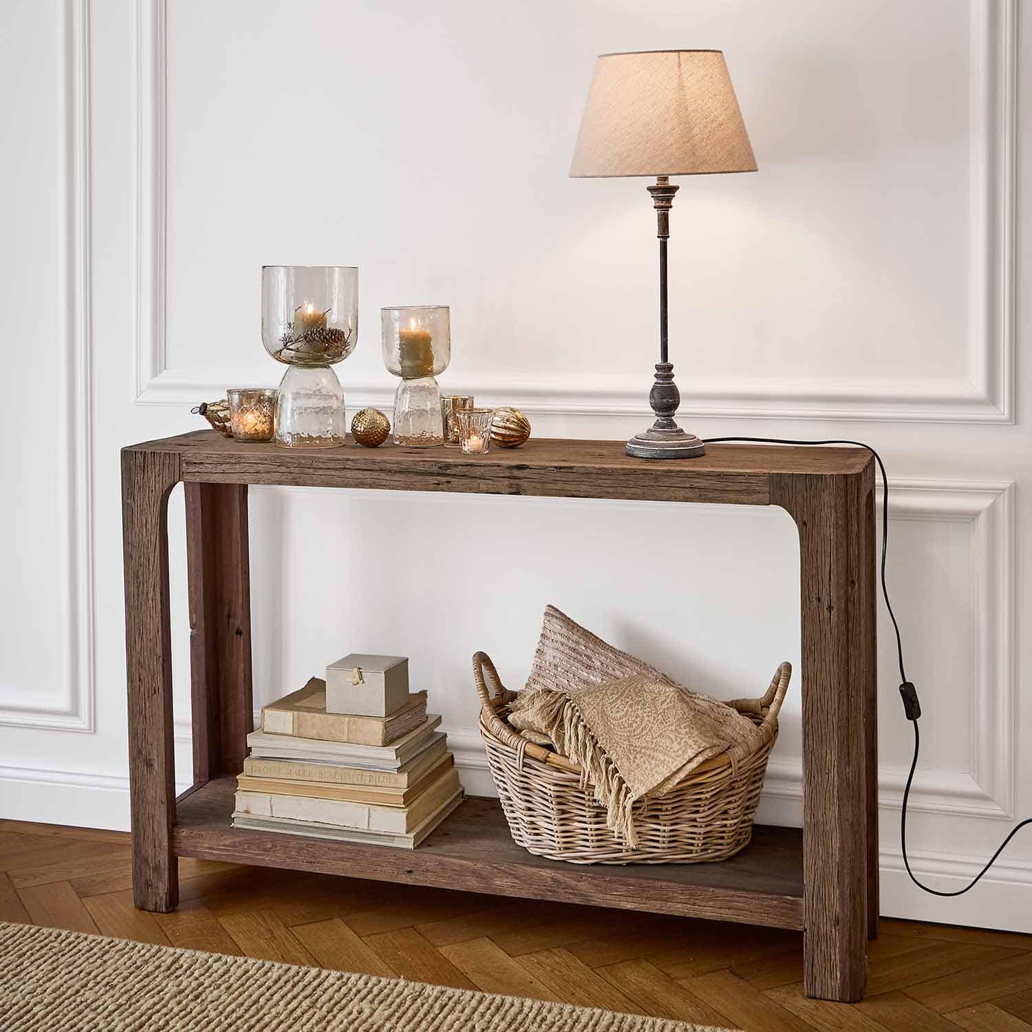 Houten sidetable met tafellamp, kaarsen in glazen houders en rieten mand met textiel