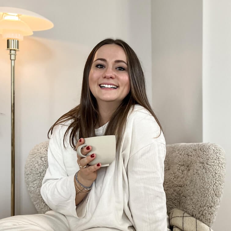 Lachende vrouw in witte trui met beige mok zittend in een comfortabele stoel in een gezellige kamer