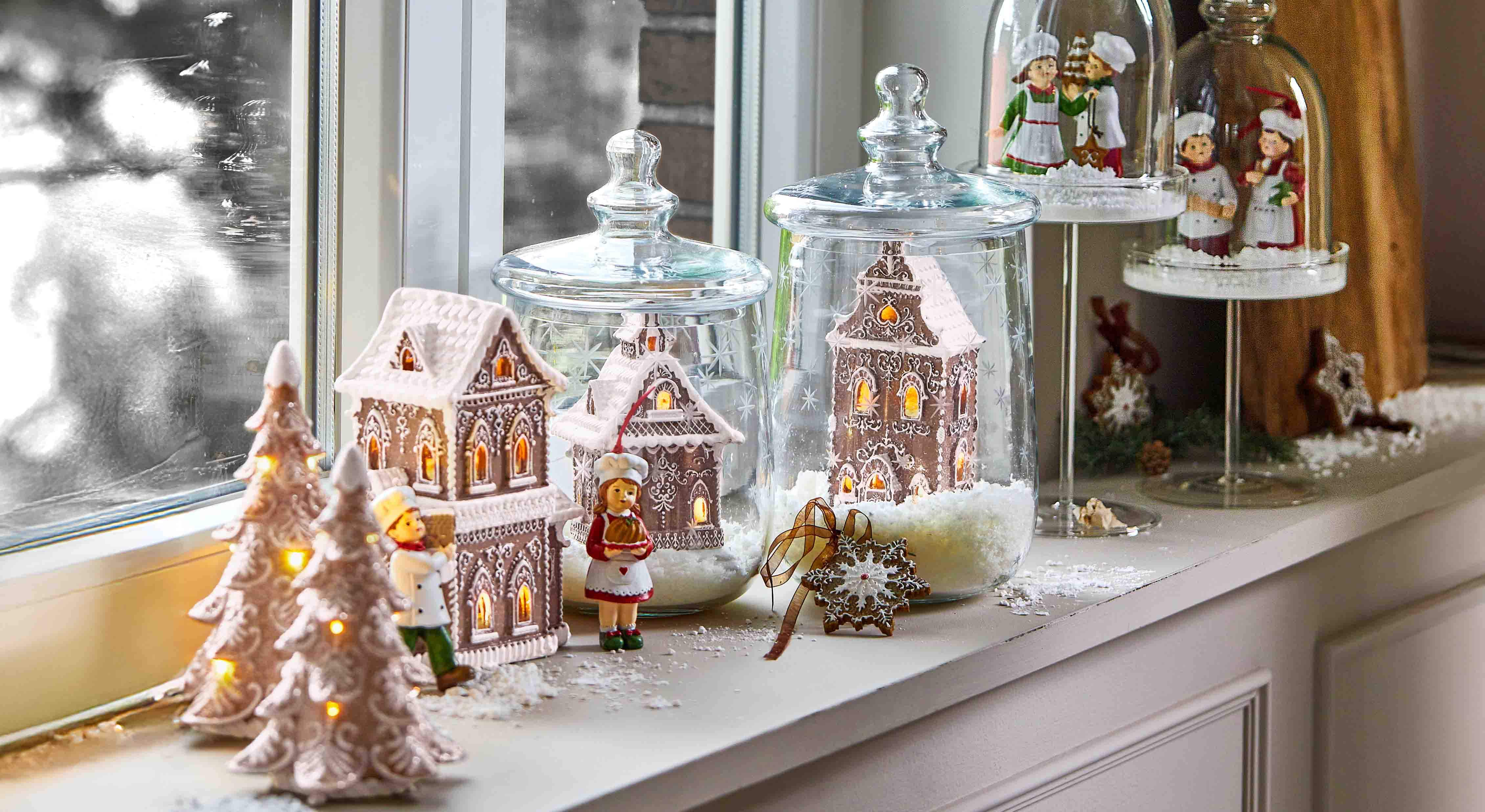 Decoratieve peperkoekhuisjes en kokfiguren onder glazen stolpen op vensterbank met winterverlichting