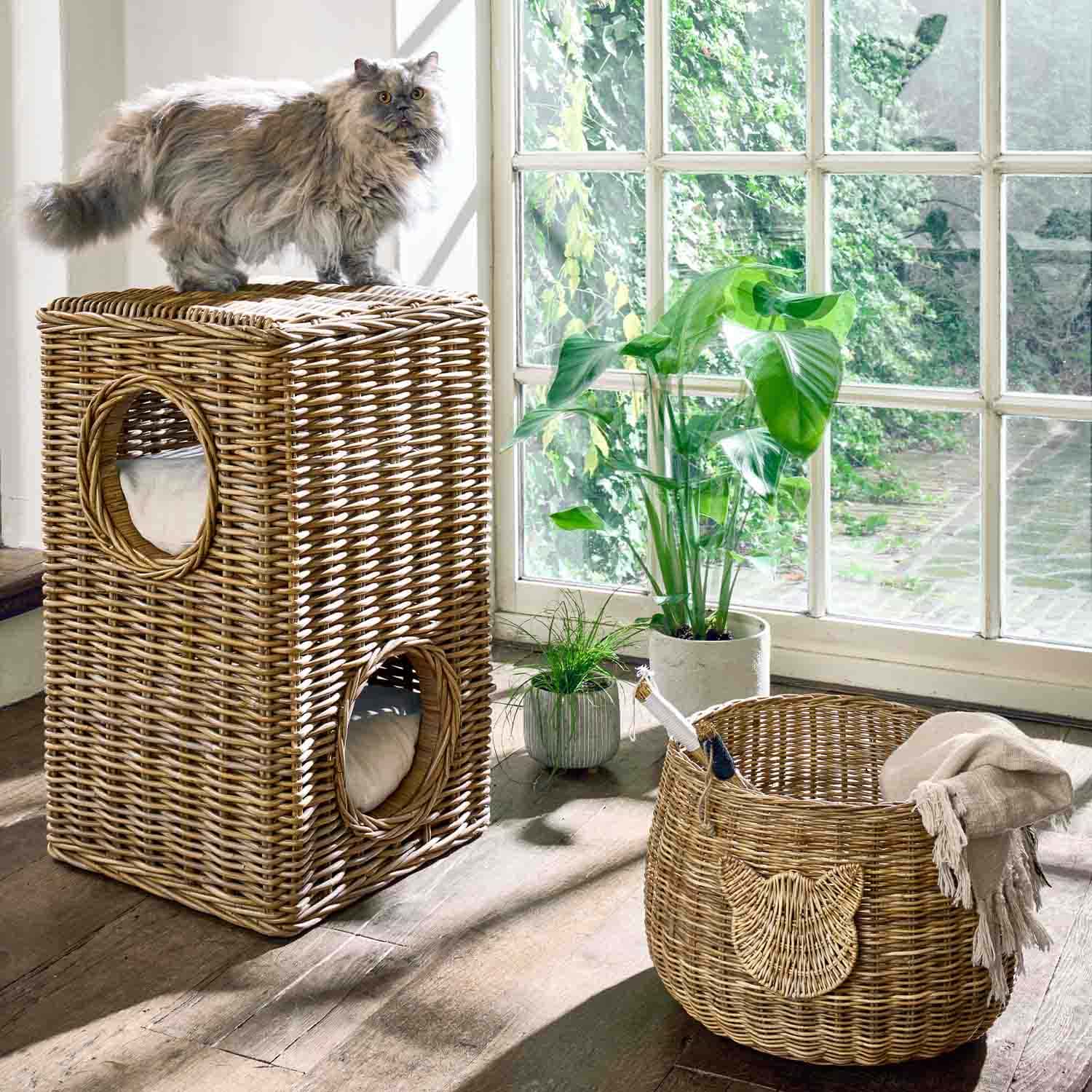 Twee verdiepingen tellend kattenhuis van natuurlijk riet met kussens naast wasmand en planten bij raam