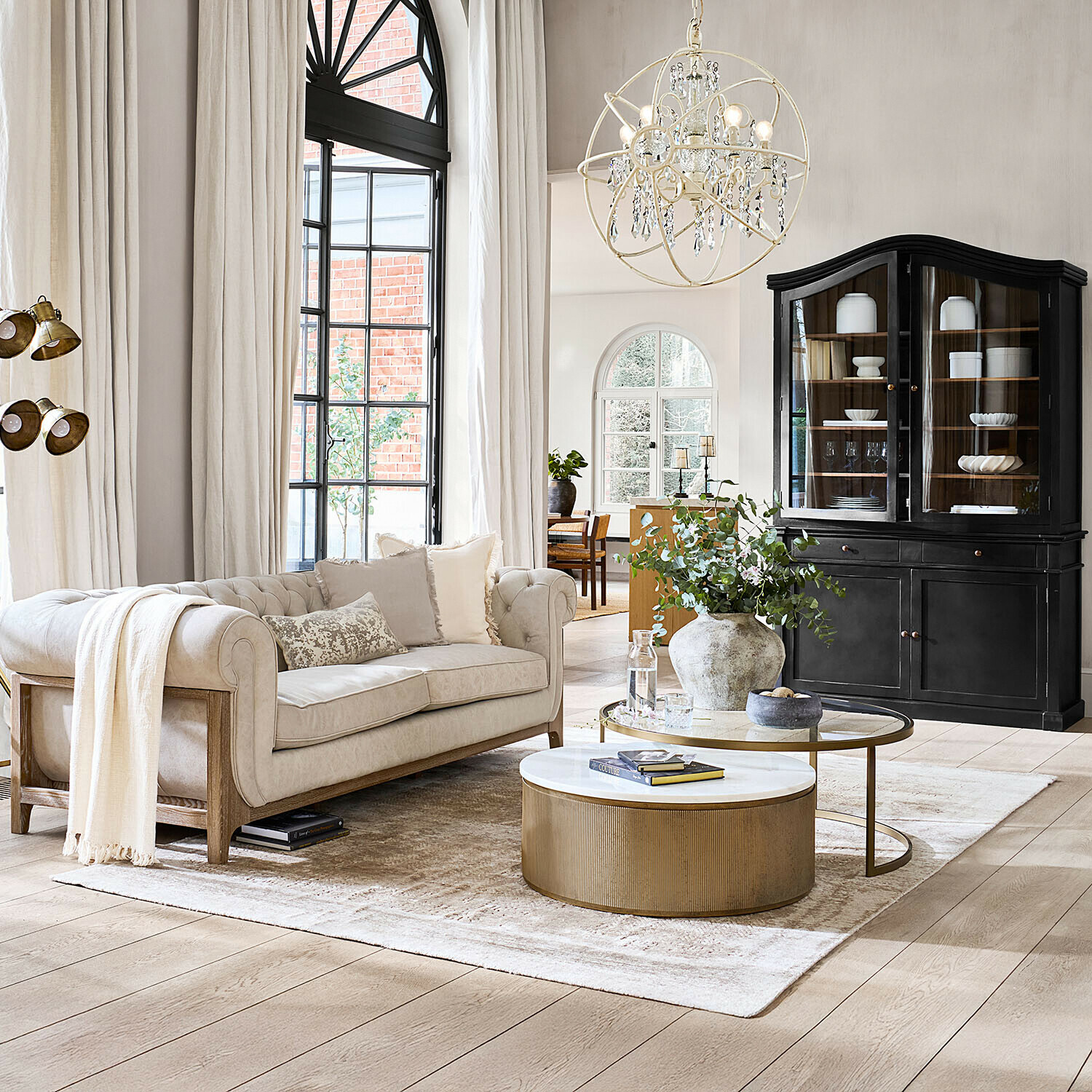 Vloerkleed Ashmere Comfortabele beige bank met houten frame in lichte woonkamer met zwarte vitrinekast en gouden salontafel