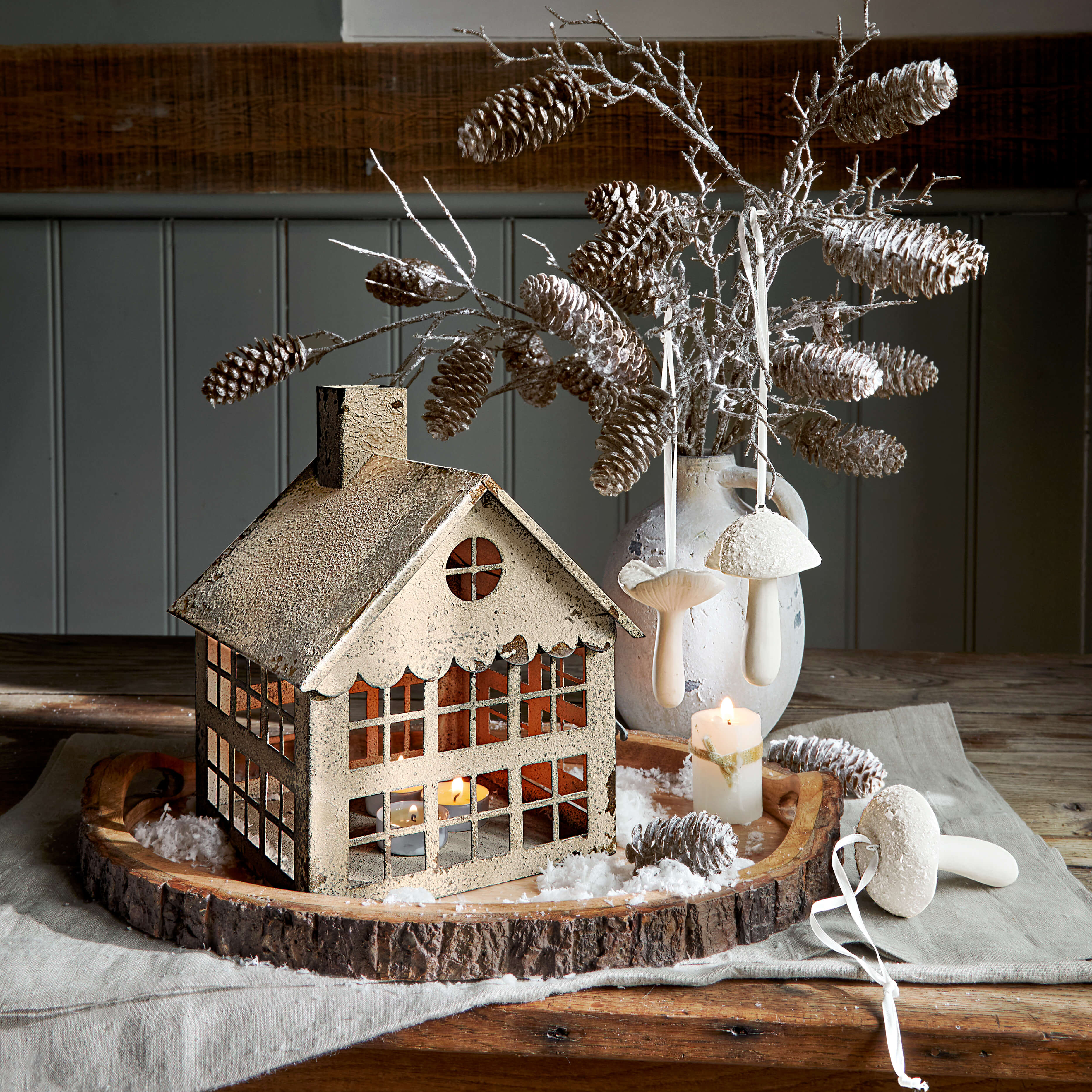 Decoratief metalen huisje met theelicht op houten schaal met dennenappels en witte paddenstoelen