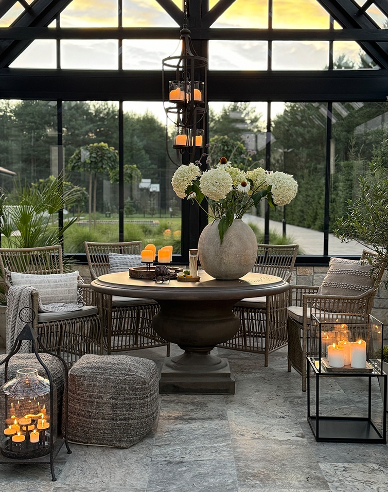 Gezellige serre met ronde tafel, rotan stoelen en decoratieve kaarsen op stenen vloer