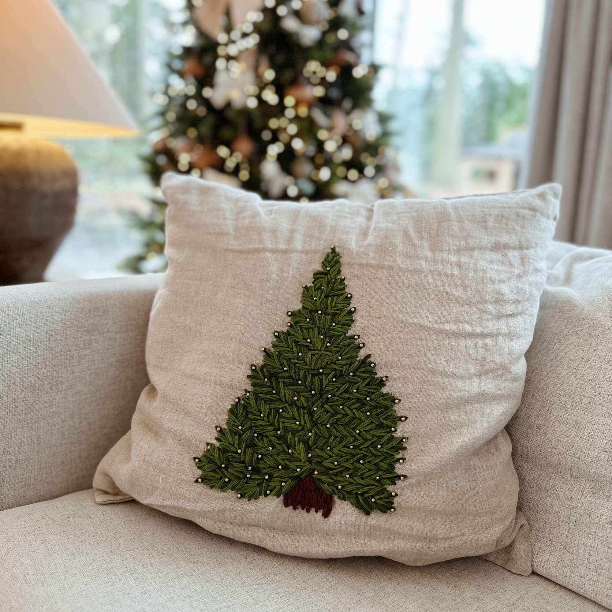 Beige linnen kussen met groene kerstboom borduurwerk en bruine stam op bank in woonkamer