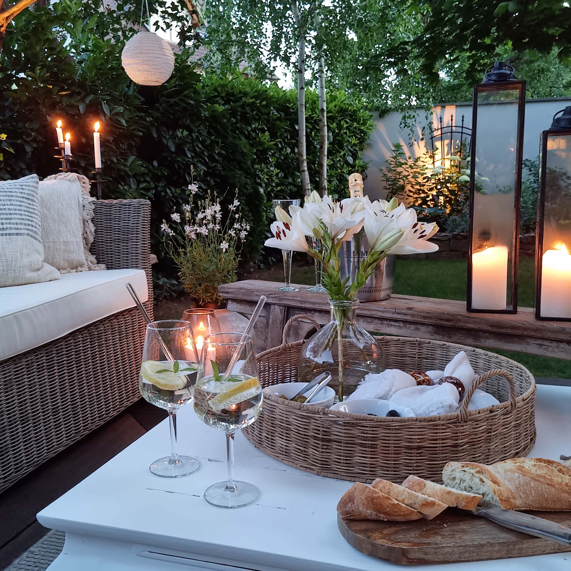 Gezellige tuin met rotan bank, witte lelies en lantaarns op witte houten tafel