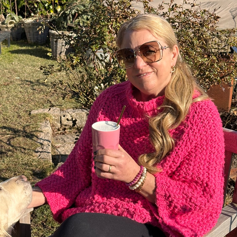 Vrouw in dikke roze trui houdt roze beker vast buiten op zonnige dag met hond naast zich