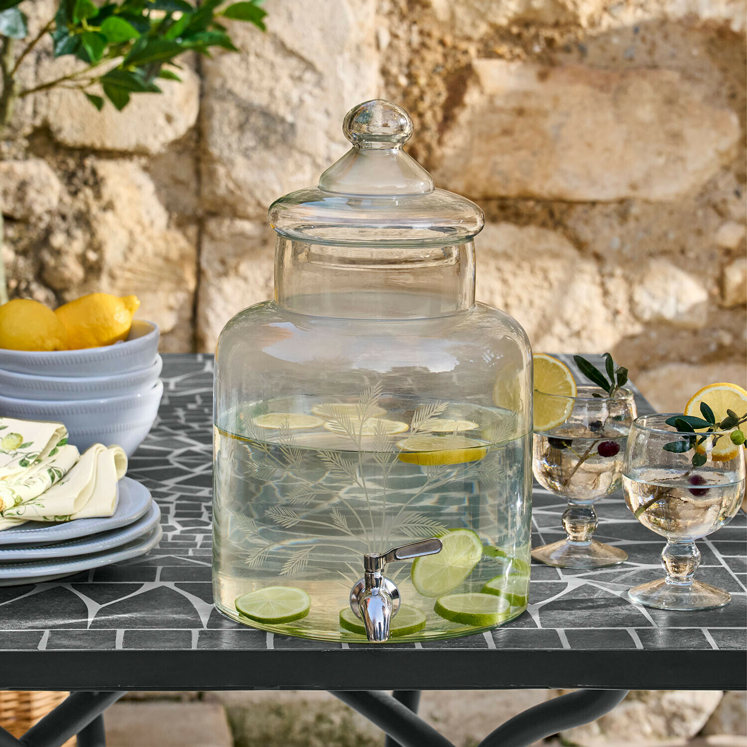 Glazen drankdispenser met deksel en kraan, gevuld met citroen en limoen, op tuintafel