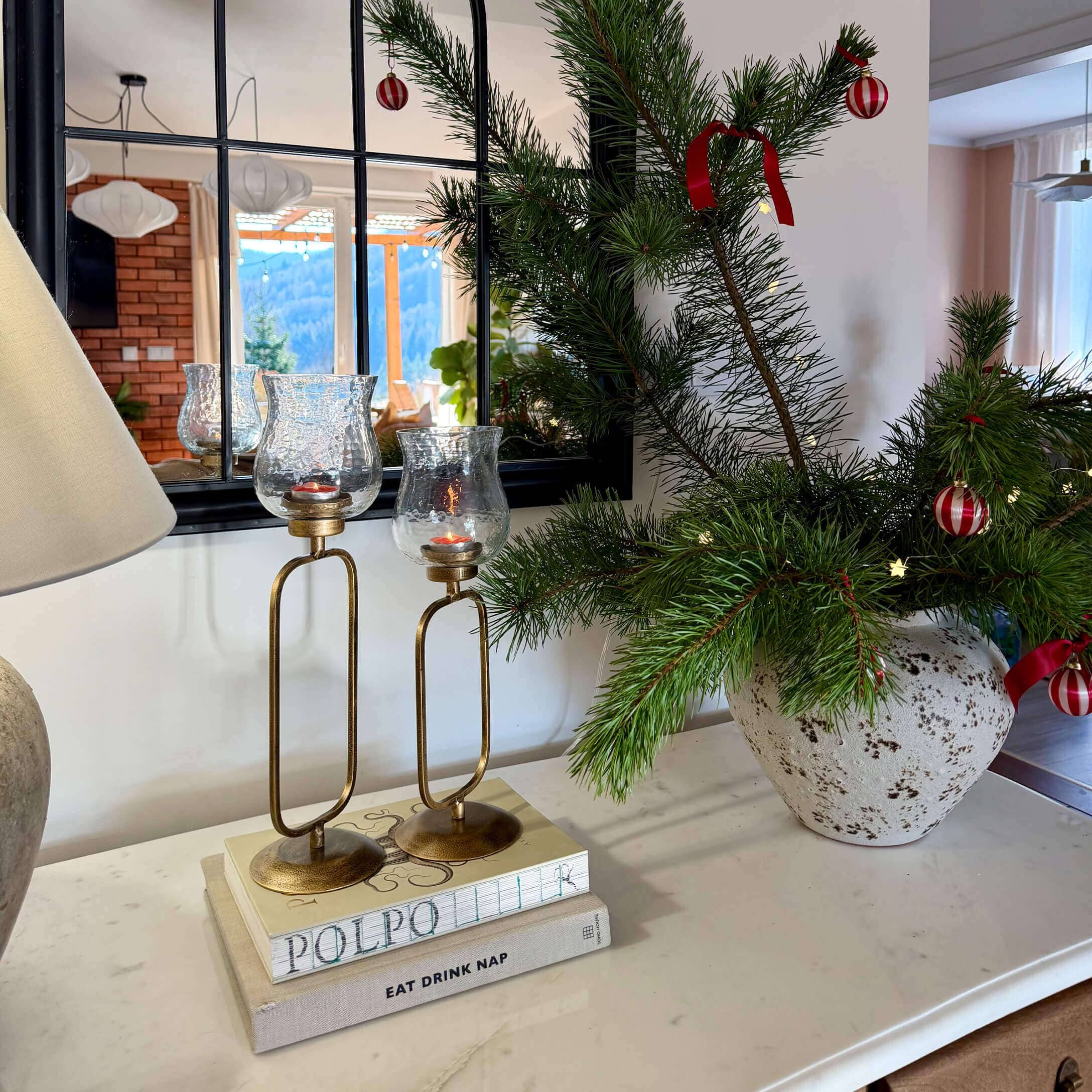 Gouden kandelaars met helder glas op boeken naast een vaas met groene takken en rode versieringen