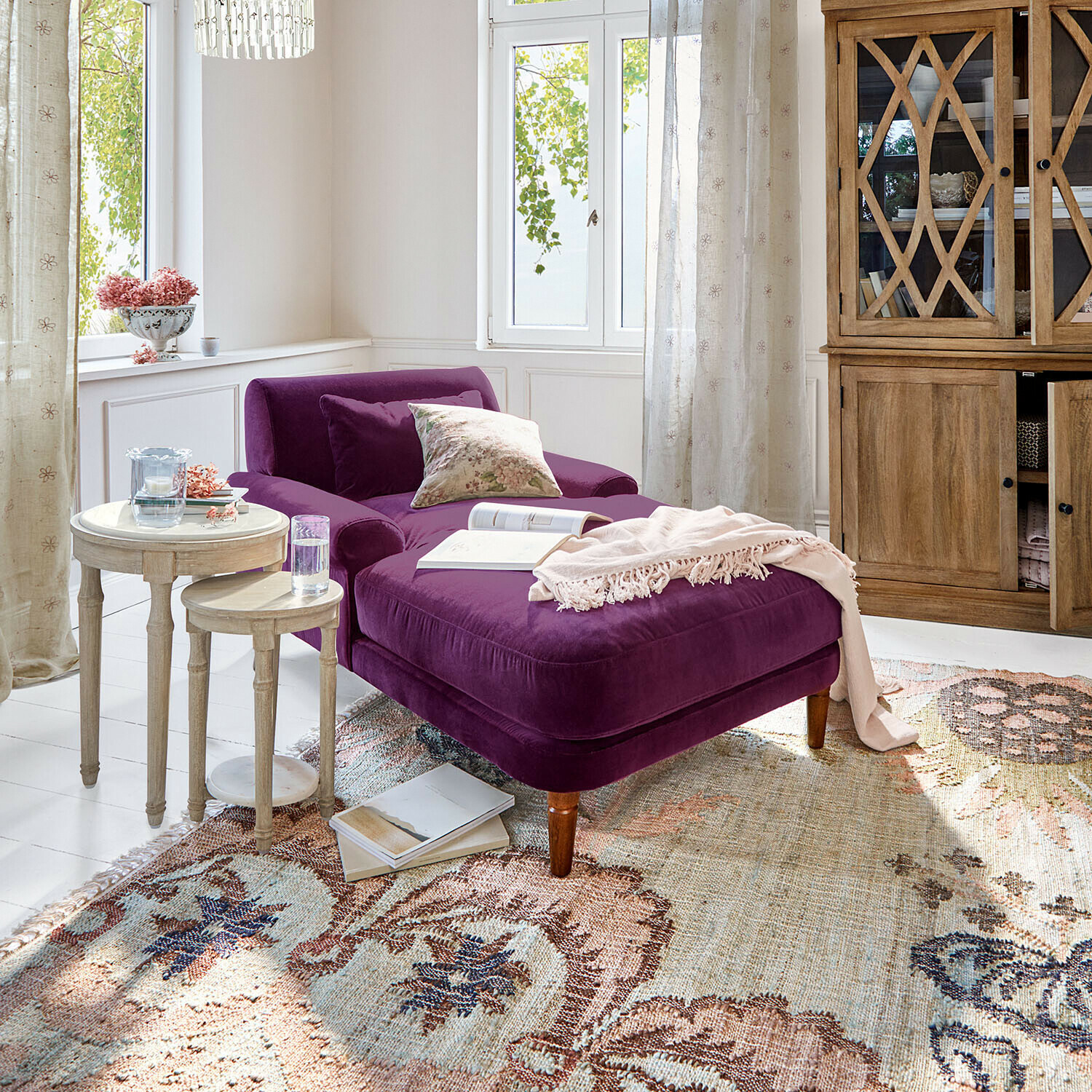 Paarse fluwelen chaise longue in lichte woonkamer met houten kast en twee ronde bijzettafels