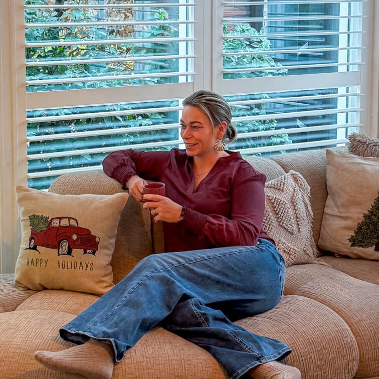 Vrouw in bordeaux blouse en spijkerbroek ontspant op beige bank met rode kerstkussen en kopje
