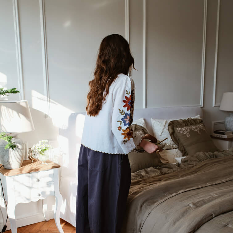 Vrouw in witte blouse met kleurrijke borduursels op mouwen in slaapkamer met bruin beddengoed en lamp op nachtkastje
