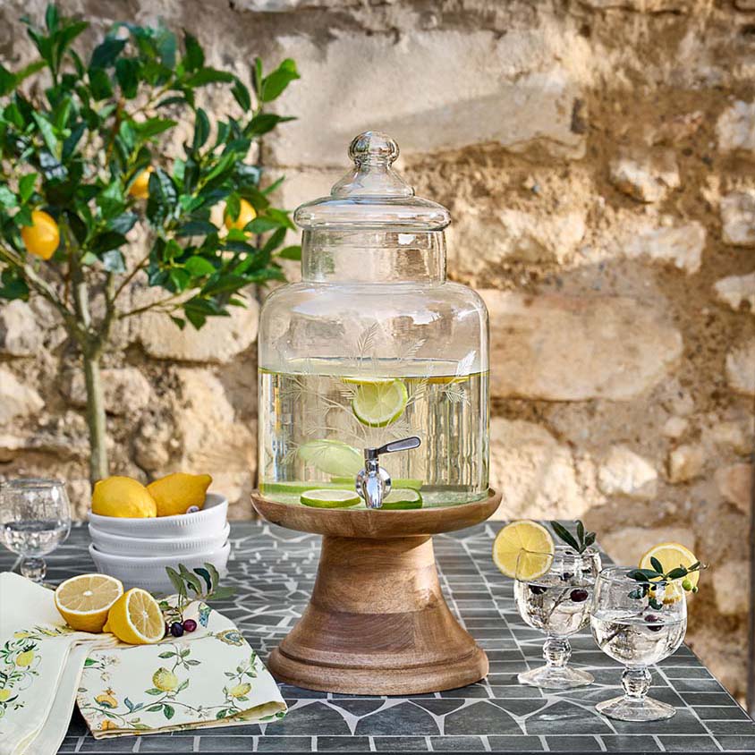 Glazen drankendispenser met deksel op houten voet, limoen- en citroenschijfjes op tafel