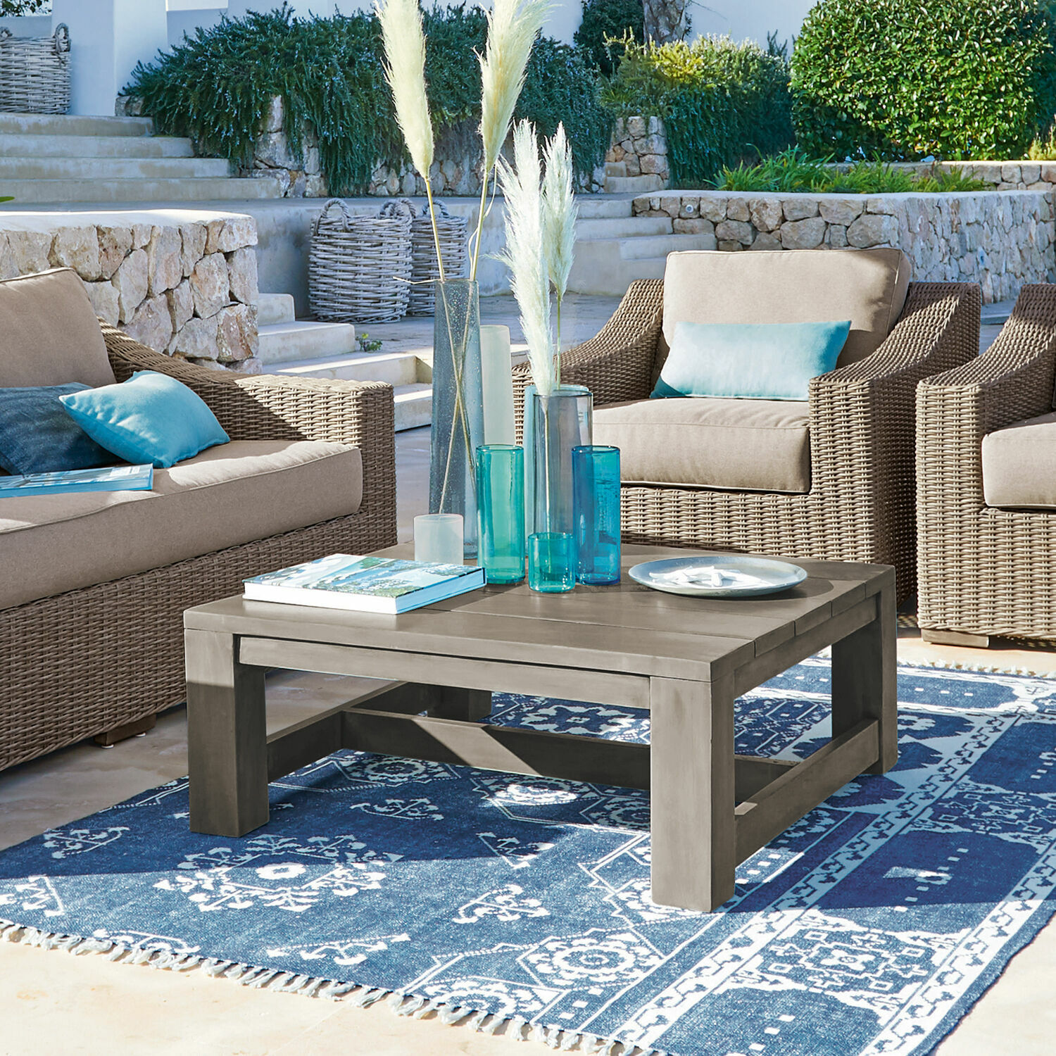 Salontafel Malden Grijze houten salontafel op blauw patroon vloerkleed, omringd door rotan stoelen met blauwe kussens.