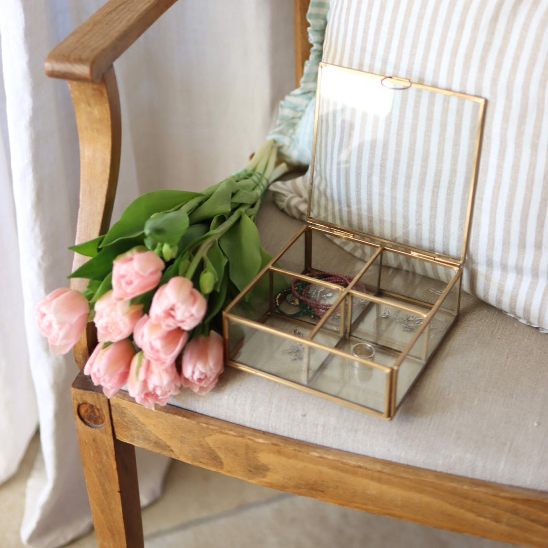 Glazen sieradendoos met messing frame op houten stoel naast roze tulpen