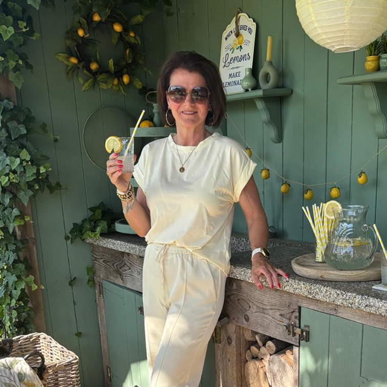 Vrouw in crèmekleurige outfit ontspant met limonade voor groene muur met citroendecoratie