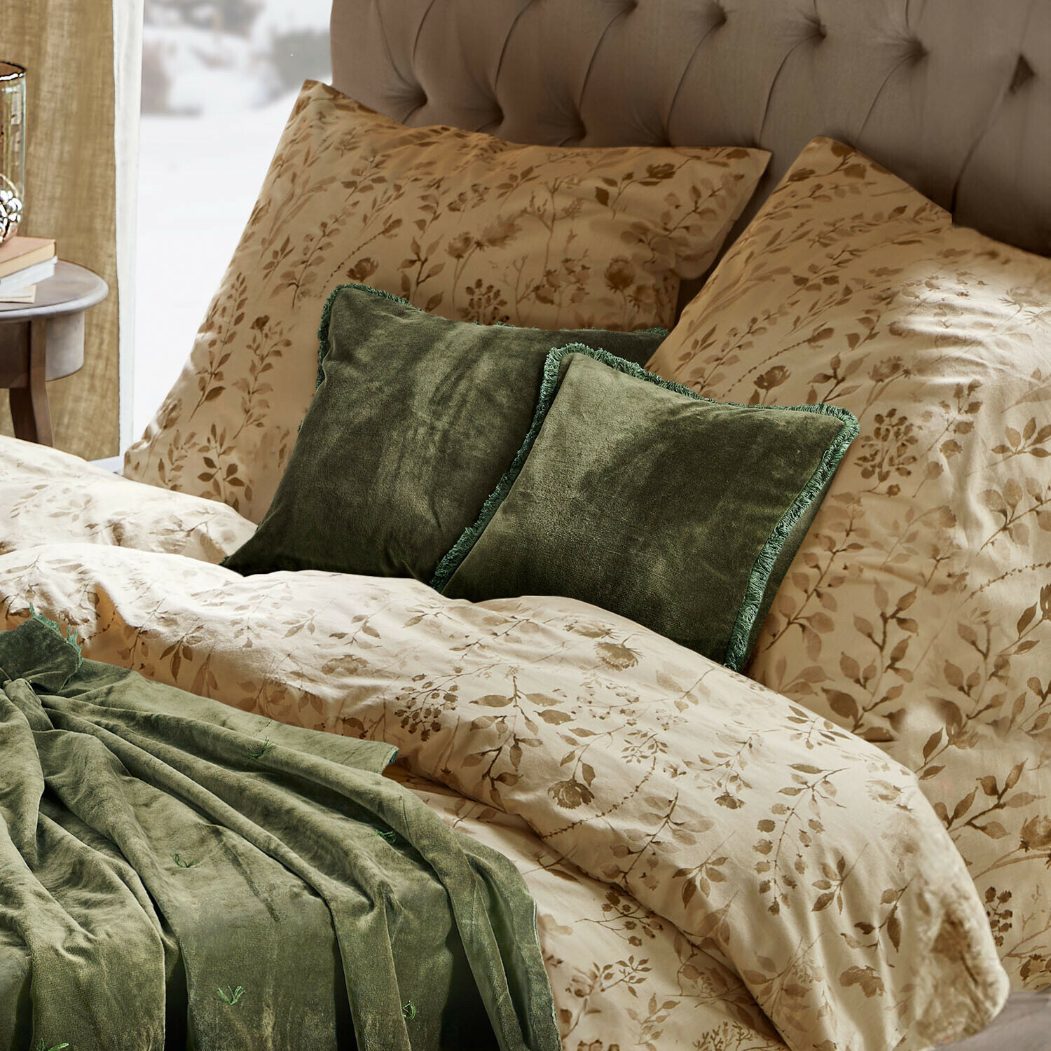 Beige beddengoed met bloemmotief, groene fluwelen kussens en deken op een gestoffeerd bed