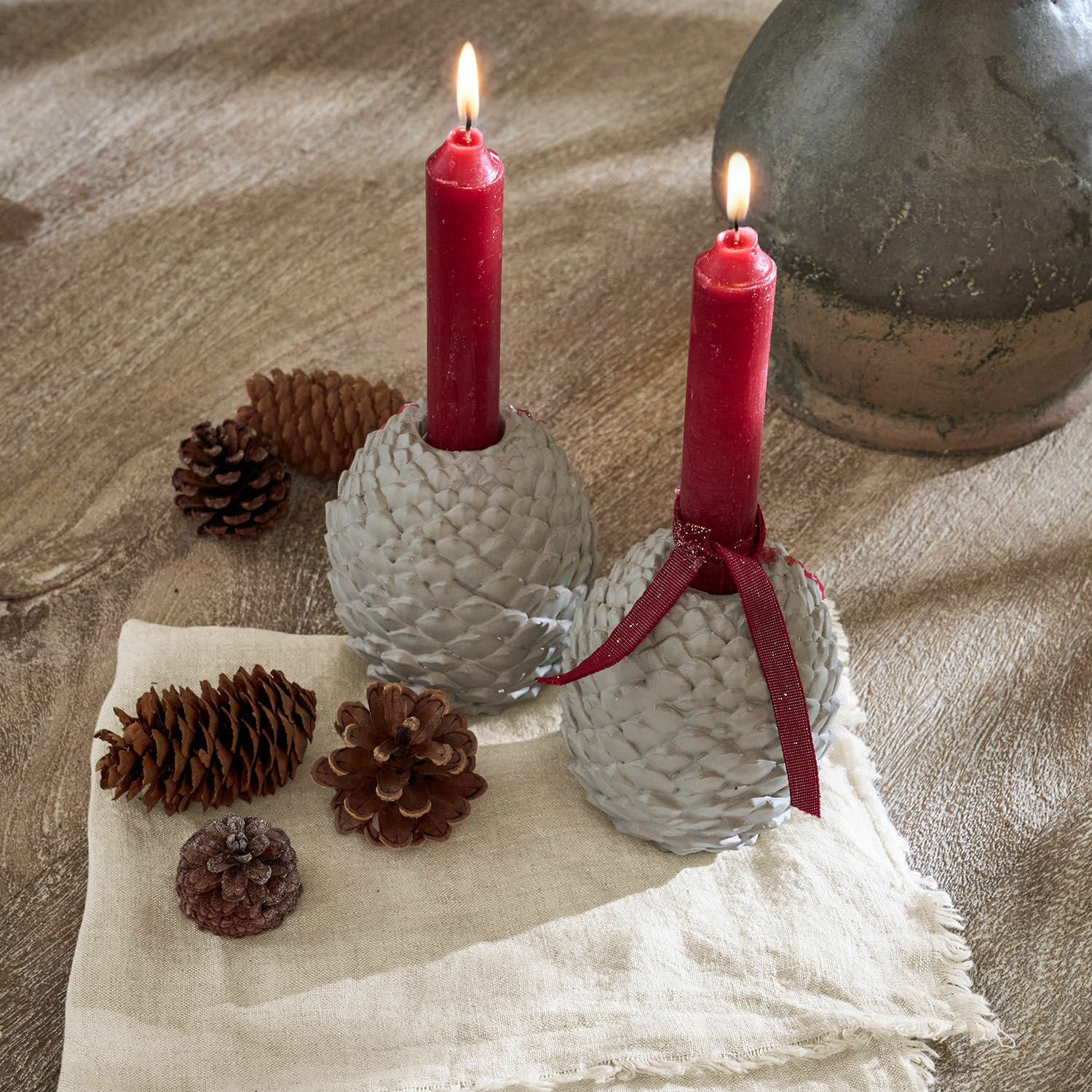 Twee grijze dennenappelkaarsenhouders met rode kaarsen op linnen doek en dennenappels