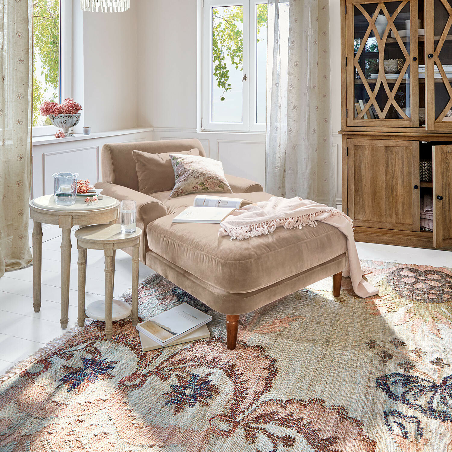 Beige fluwelen chaise longue met kussens en roze deken op een patroon vloerkleed in lichte woonkamer