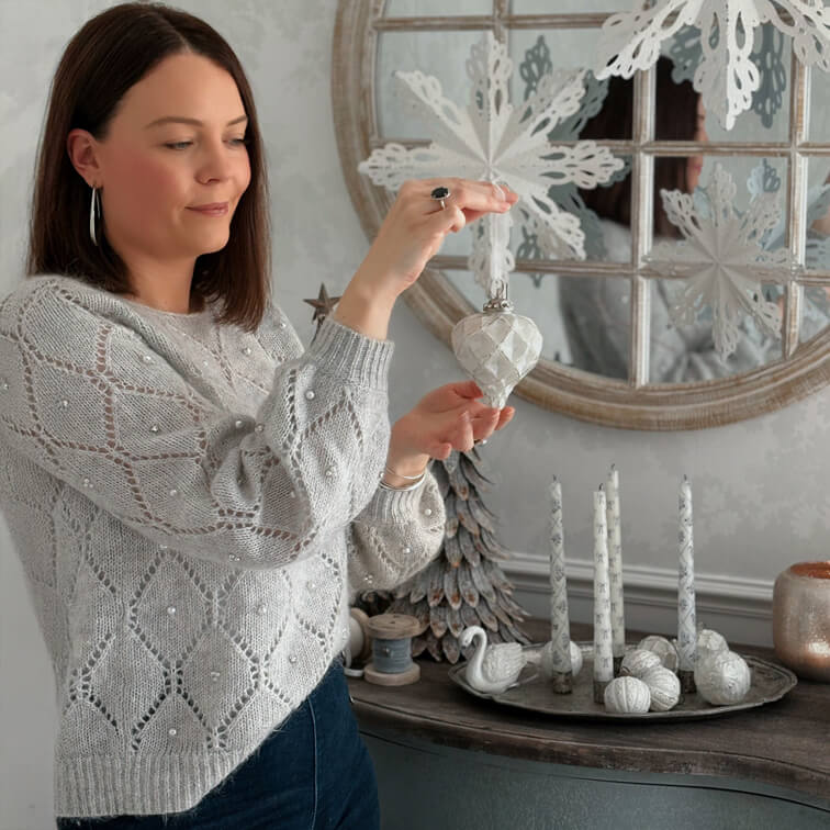 Vrouw in grijze trui met parels houdt witte kerstversiering vast in gezellige ruimte met winterdecoratie