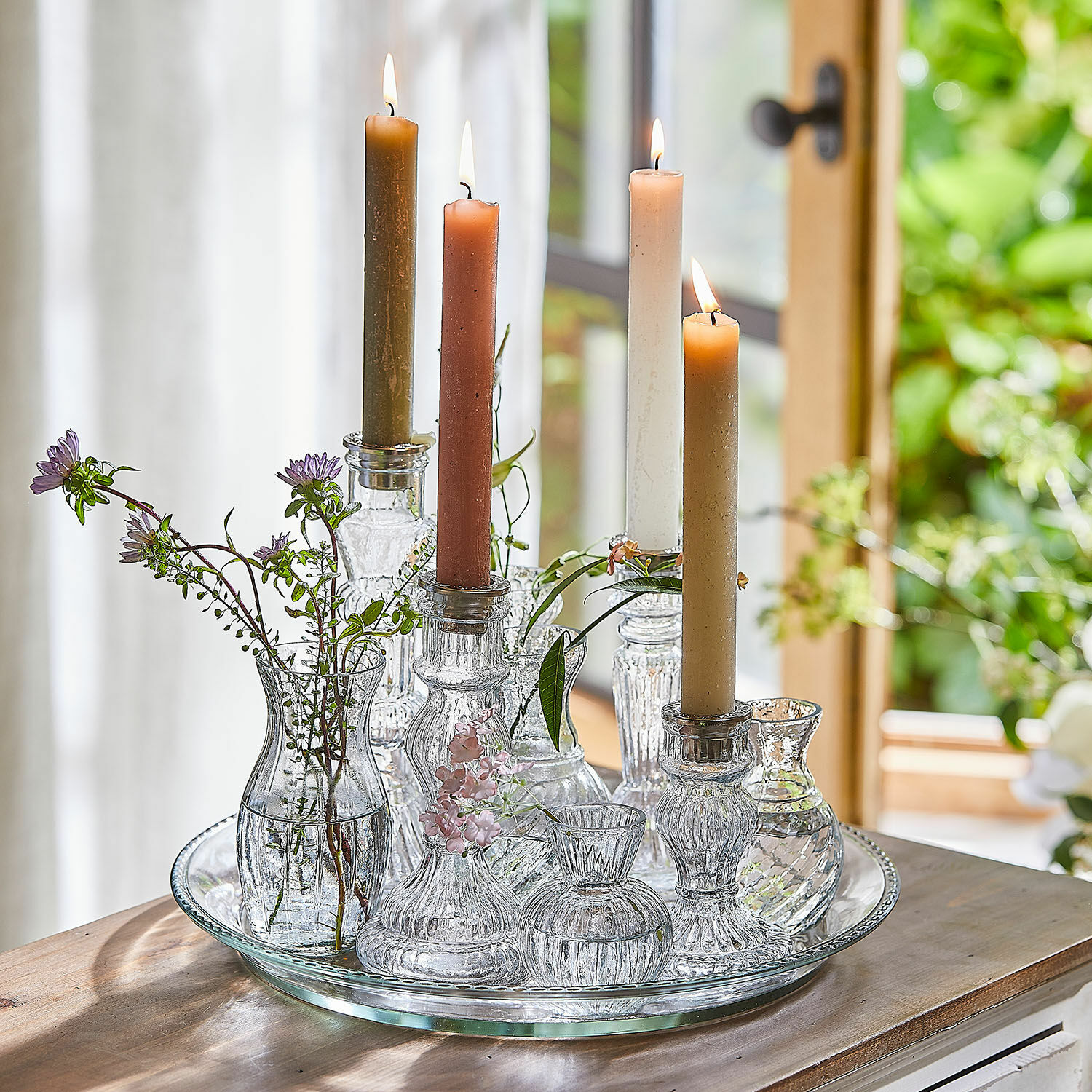 Kaars set van 12 Bauceau Glazen kandelaren en vaasjes met gekleurde kaarsen en bloemen op glazen dienblad op houten tafel