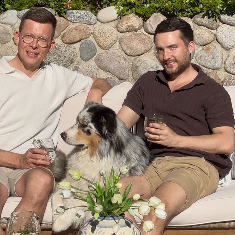 Twee mannen zitten buiten op een bank met een Australische herdershond en een vaas met witte tulpen.