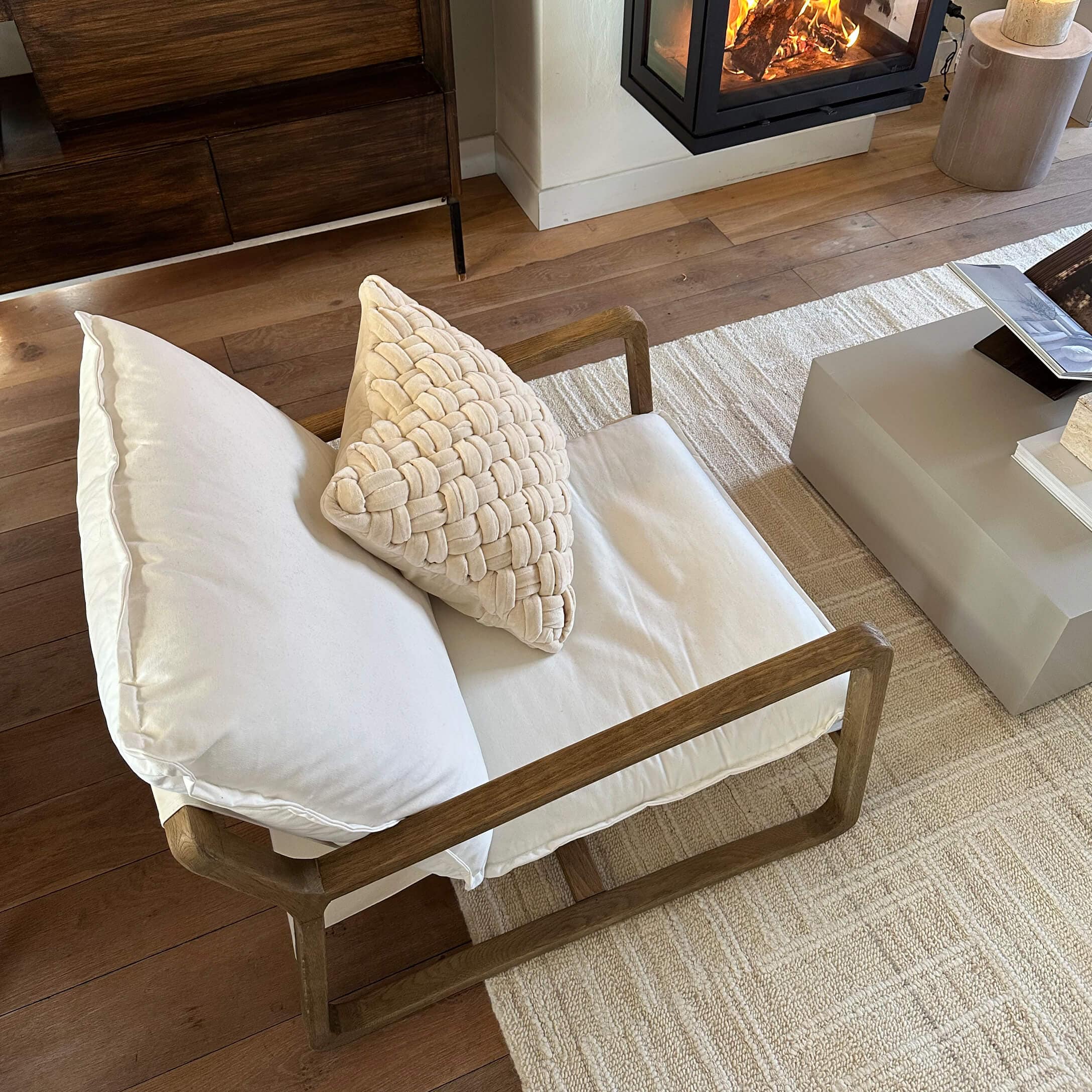 Stoel met houten frame en crèmekleurige kussens op lichte vloerkleed bij open haard in woonkamer