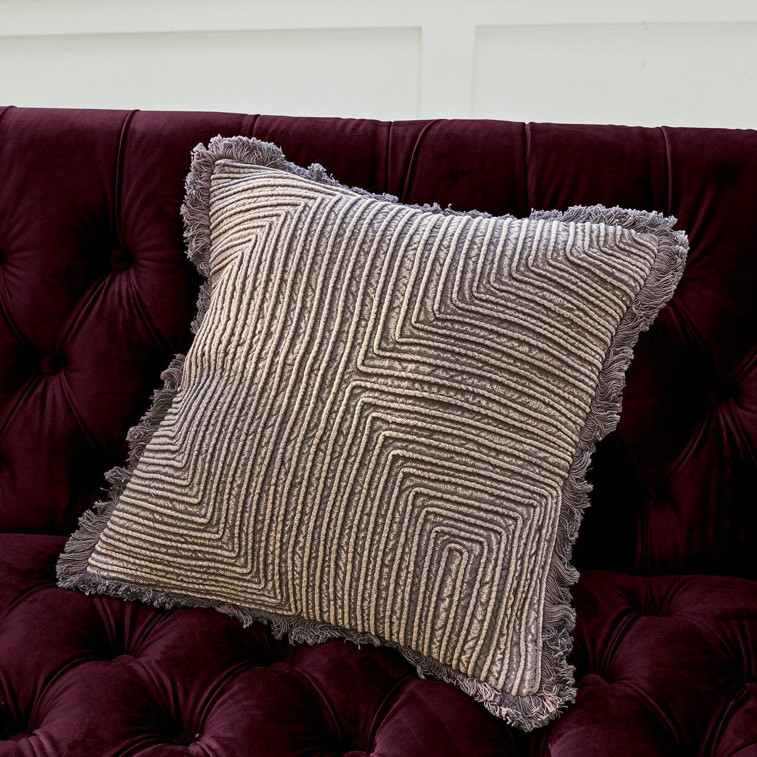 Grijs sierkussen met geometrisch patroon en franjes op bordeauxrode fluwelen bank