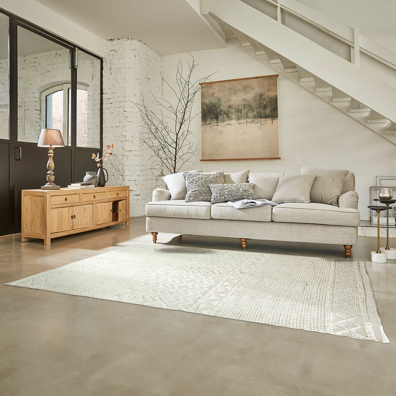 Beige bank met kussens en lichte houten dressoir in lichte woonkamer met vloerkleed en wanddecoratie