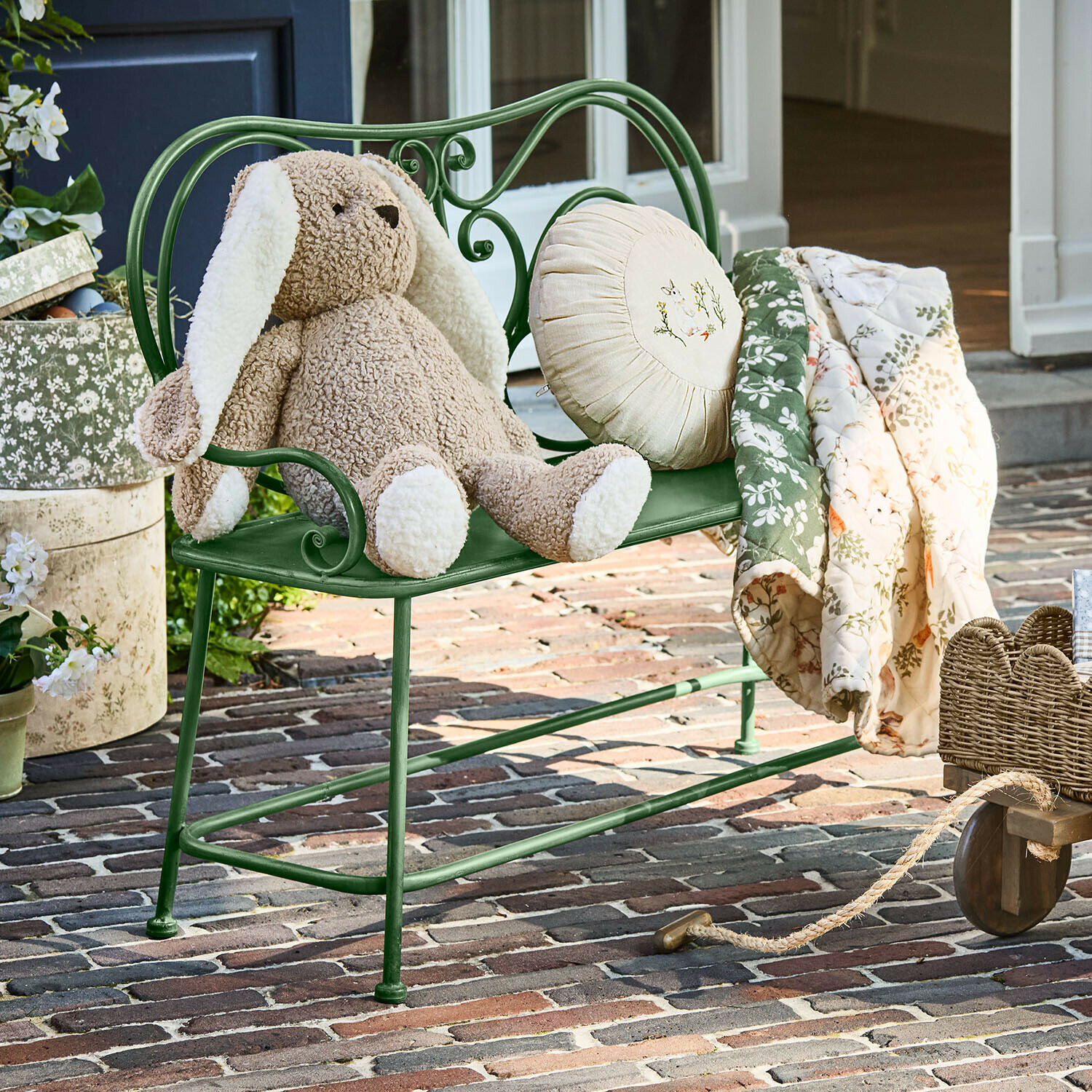 Groene metalen bank met beige pluchen konijn en bloemen deken op een bakstenen terras