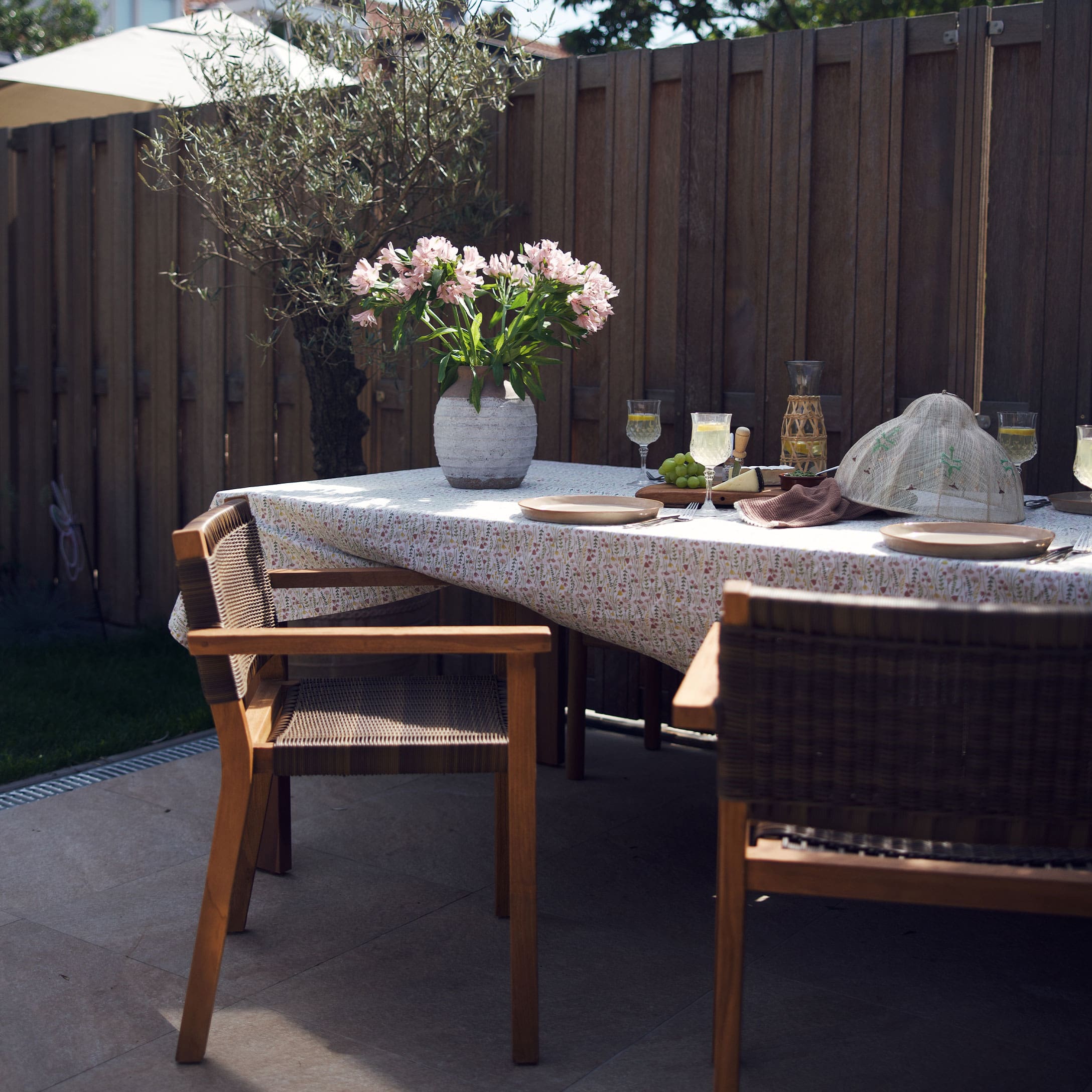 Houten tuintafel met stoelen, stenen vaas met roze bloemen op een tafelkleed met bloemenpatroon