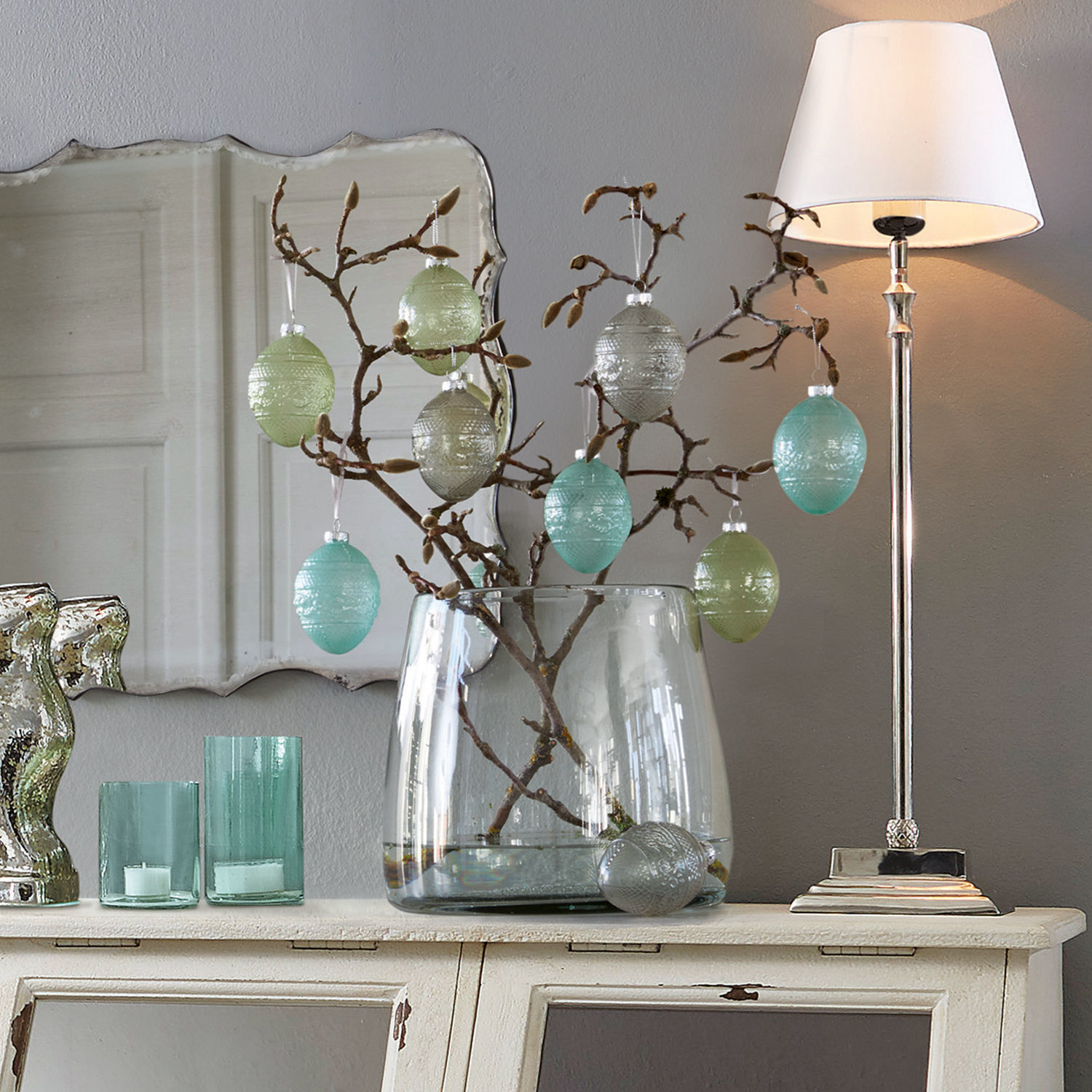 Decoratieve glazen eieren in pastelkleuren aan tak in doorzichtige vaas op witte dressoir