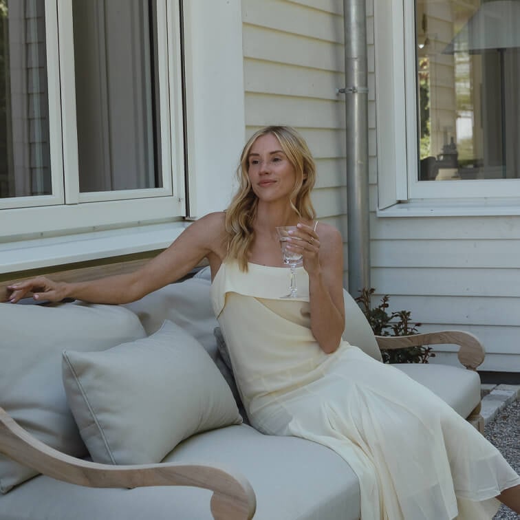 Vrouw in crèmekleurige jurk zit ontspannen op lichtgrijze bank buiten en houdt een wijnglas vast