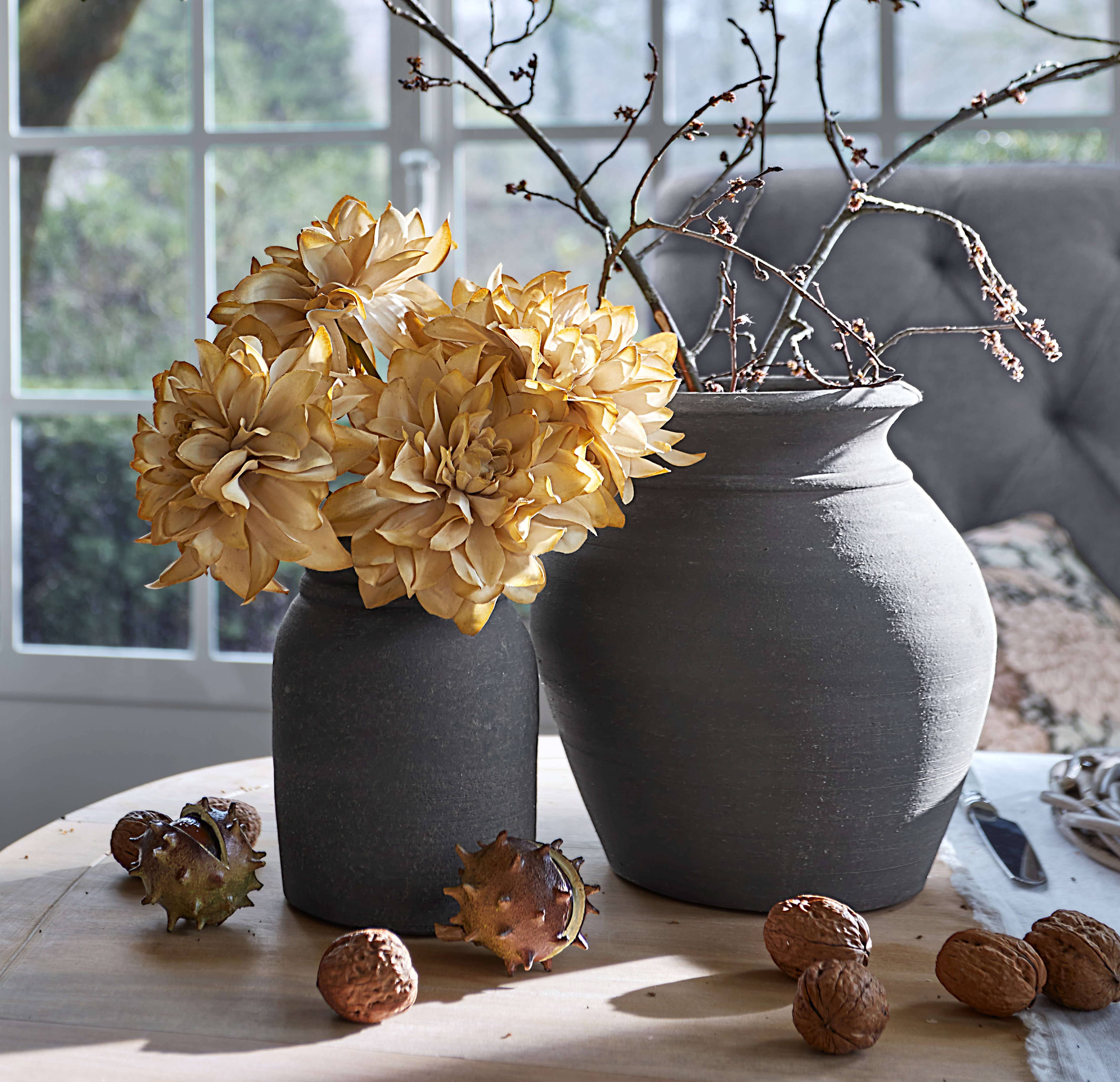 Twee grijze keramische vazen met gedroogde bloemen en takken op houten tafel met walnoten