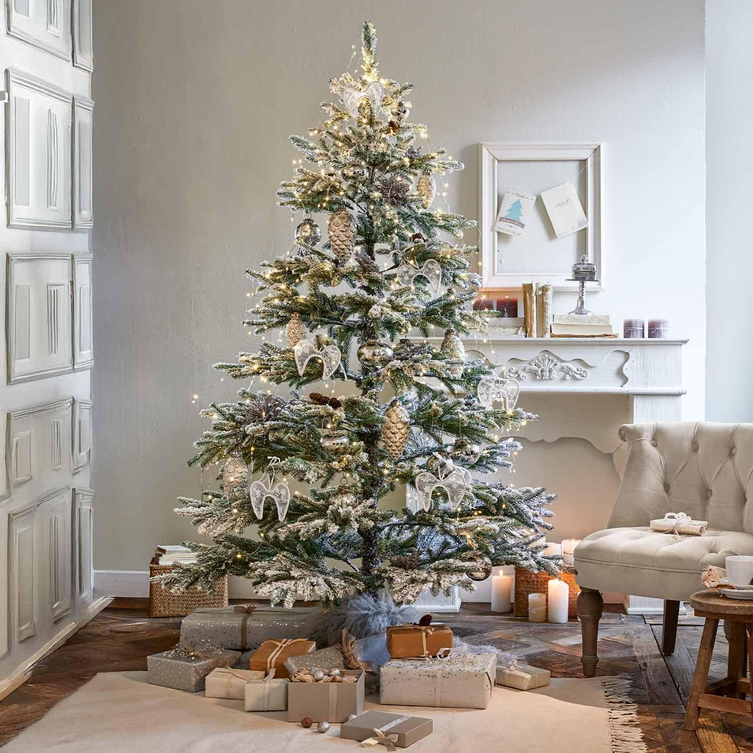 Besneeuwde kerstboom met witte engelornamenten en gouden dennenappels in een gezellige woonkamer met open haard