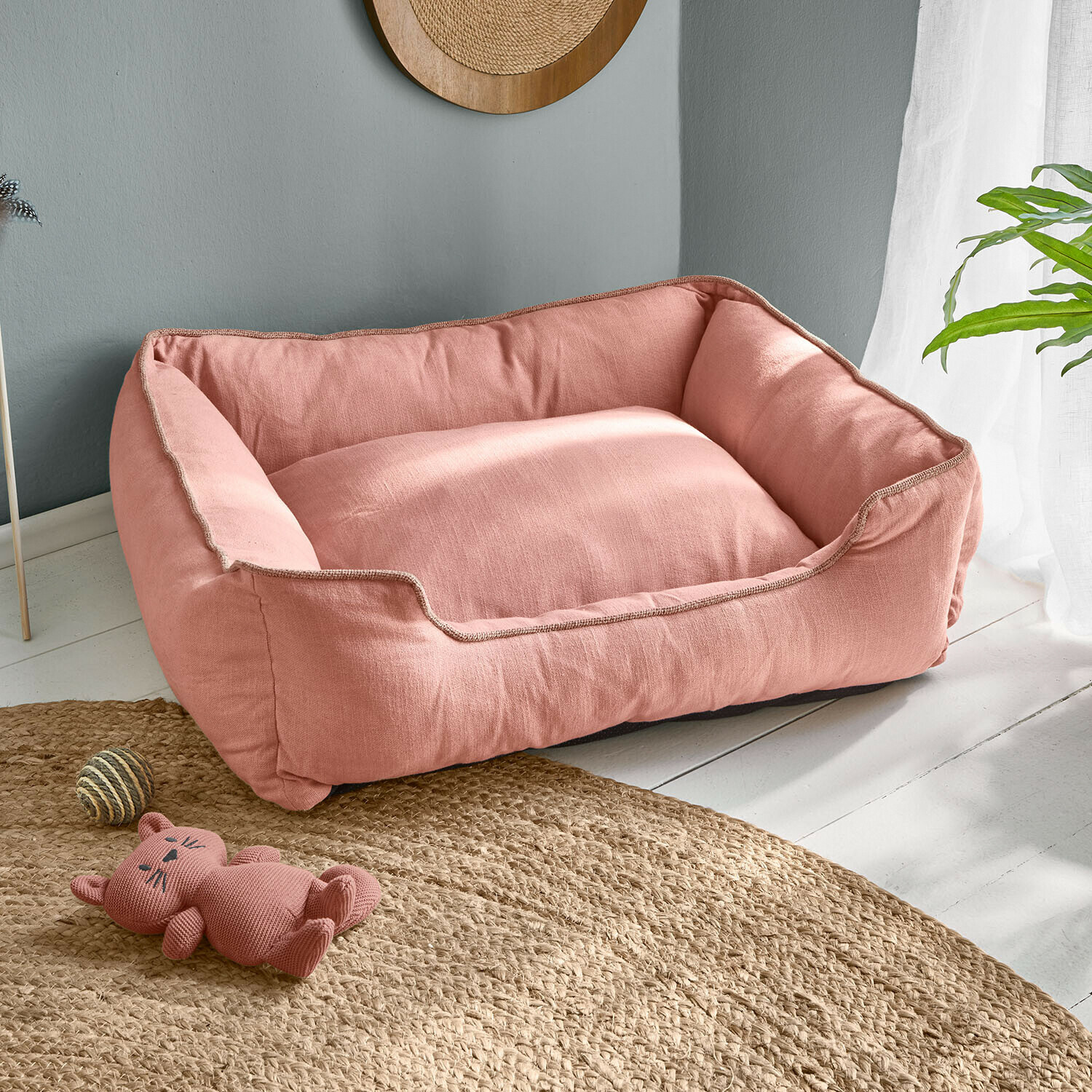 Roze hondenmand van zacht materiaal op lichte vloer met kattenknuffel en bal