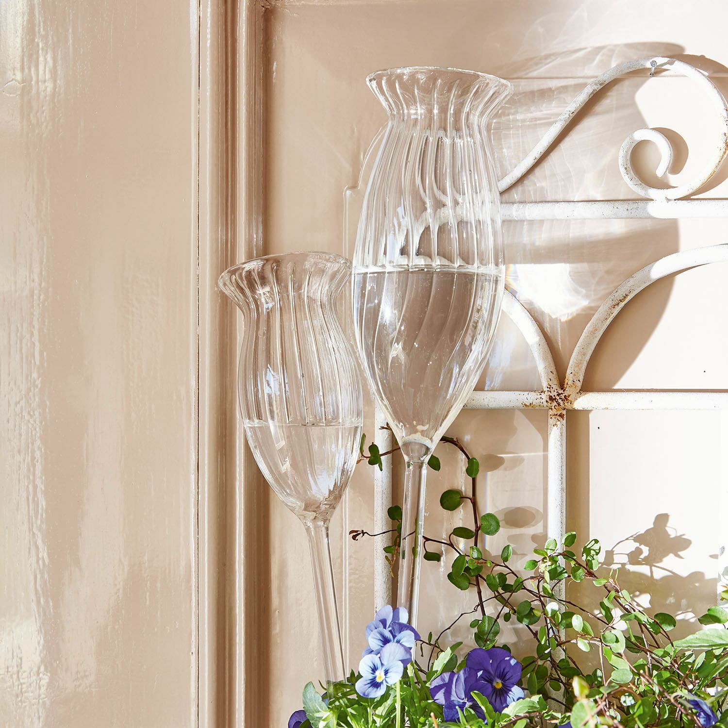 Waterdruppelaar set van 2 Radisa Tuinornamenten van helder glas met water en paarse bloemen voor een beige muur