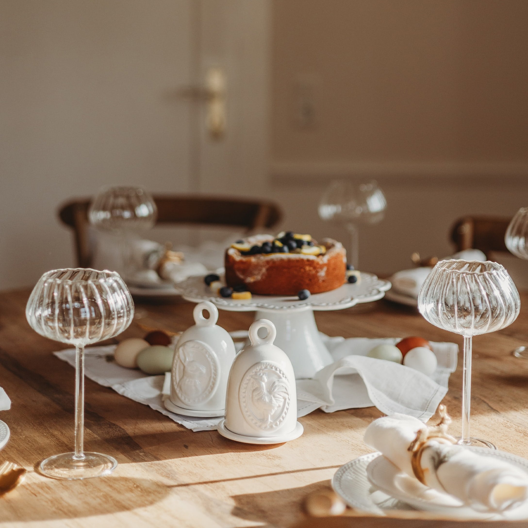 Decoratieve witte keramische klokjes met haanmotief op houten tafel met taart en glazen