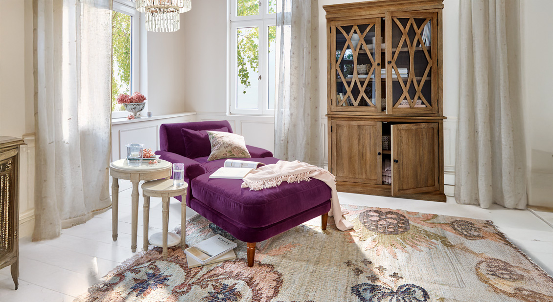 Paars fluwelen ligstoel met kussens en deken in lichte woonkamer met houten kast en vloerkleed