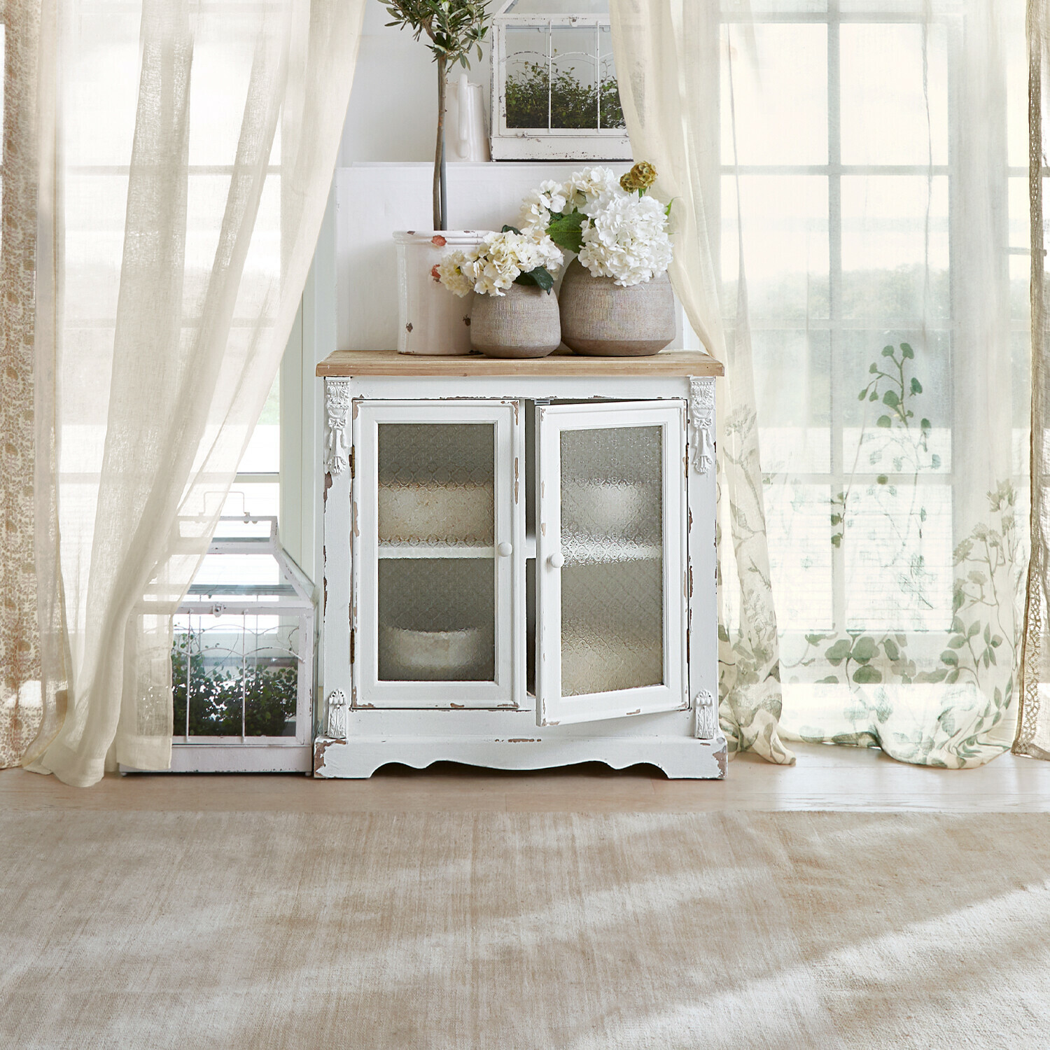 Witte houten kast met glazen deuren en decoratieve vazen met bloemen in lichte kamer