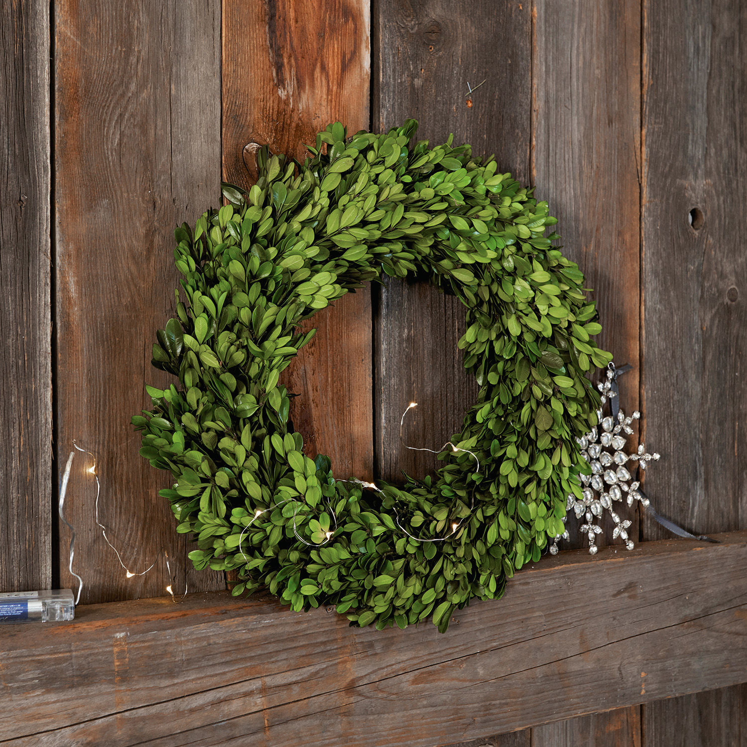 Krans Myrcella Groene buxuskrans tegen houten achtergrond met LED-lichtjes en glinsterende sneeuwvlokdecoratie