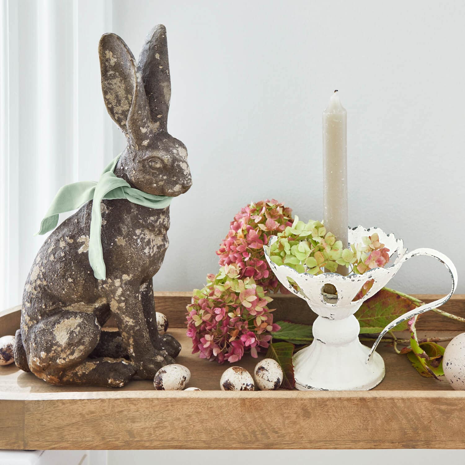 Decoratief konijn van gepatineerd metaal met groene strik, witte metalen kandelaar en bloemen op houten dienblad