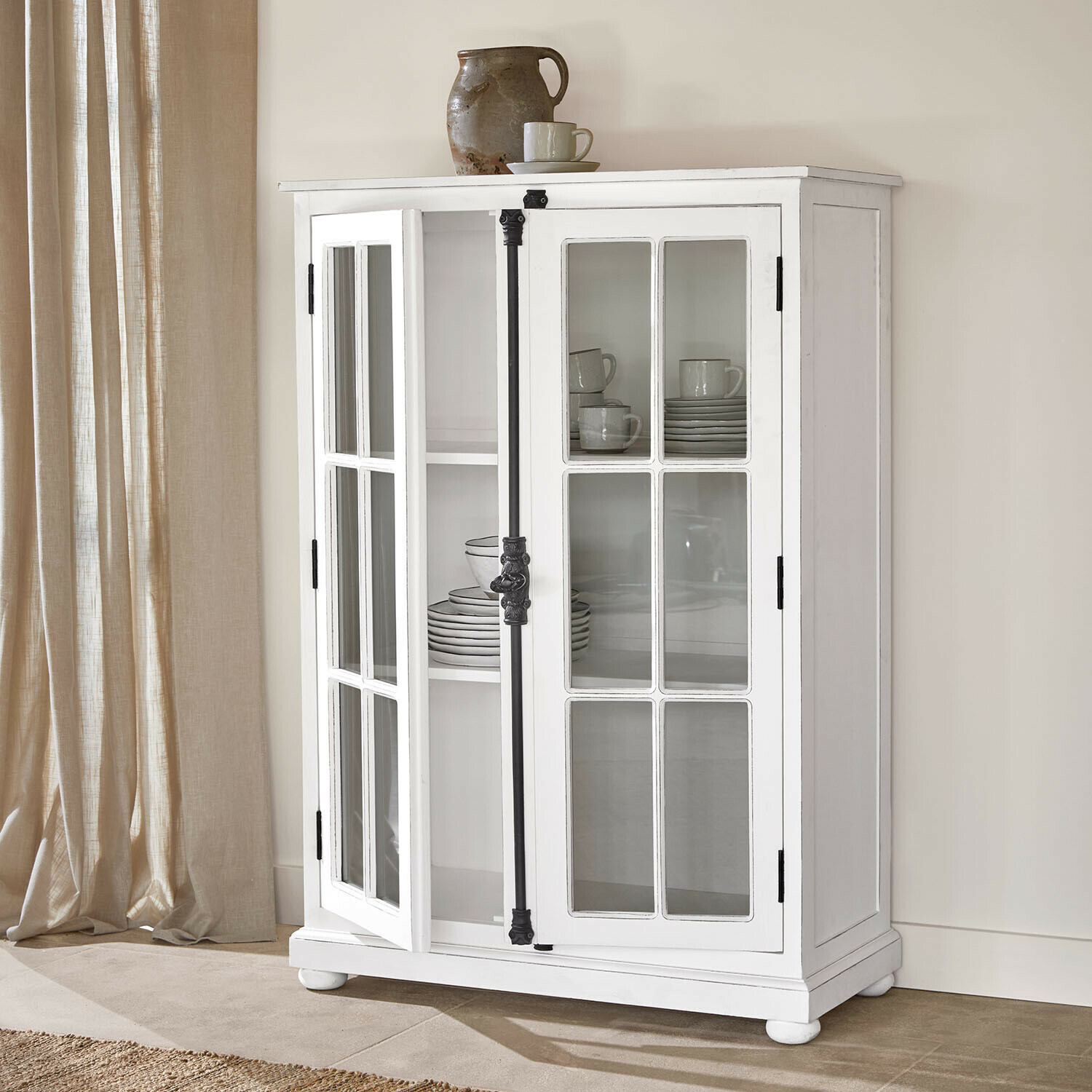 Kast Salivonne Witte houten vitrinekast met glazen deuren en zwarte metalen vintage handgrepen in lichte kamer