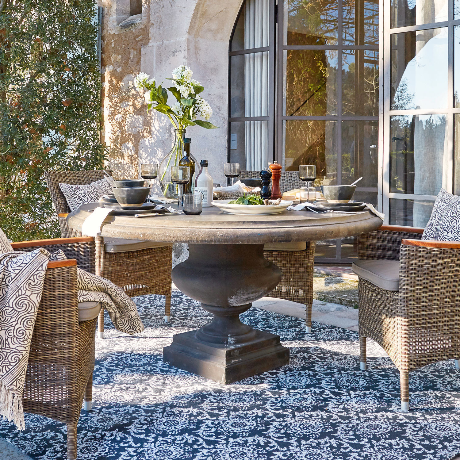 Tafel Monterey Ronde stenen tafel met zwarte voet en rieten stoelen op terras met decoratief vloerkleed