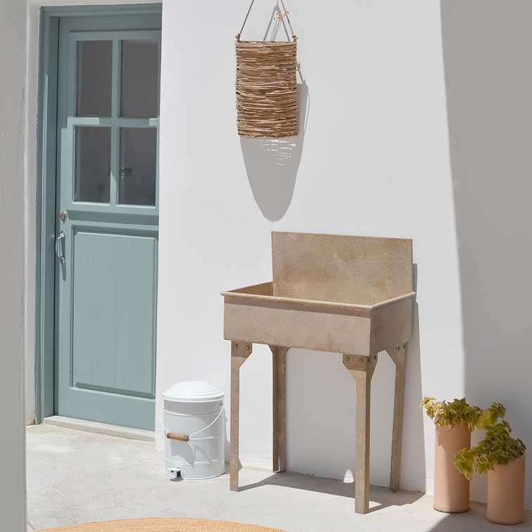 Beige stenen plantenbak op poten bij witte muur met blauwe deur en hangende rieten mand